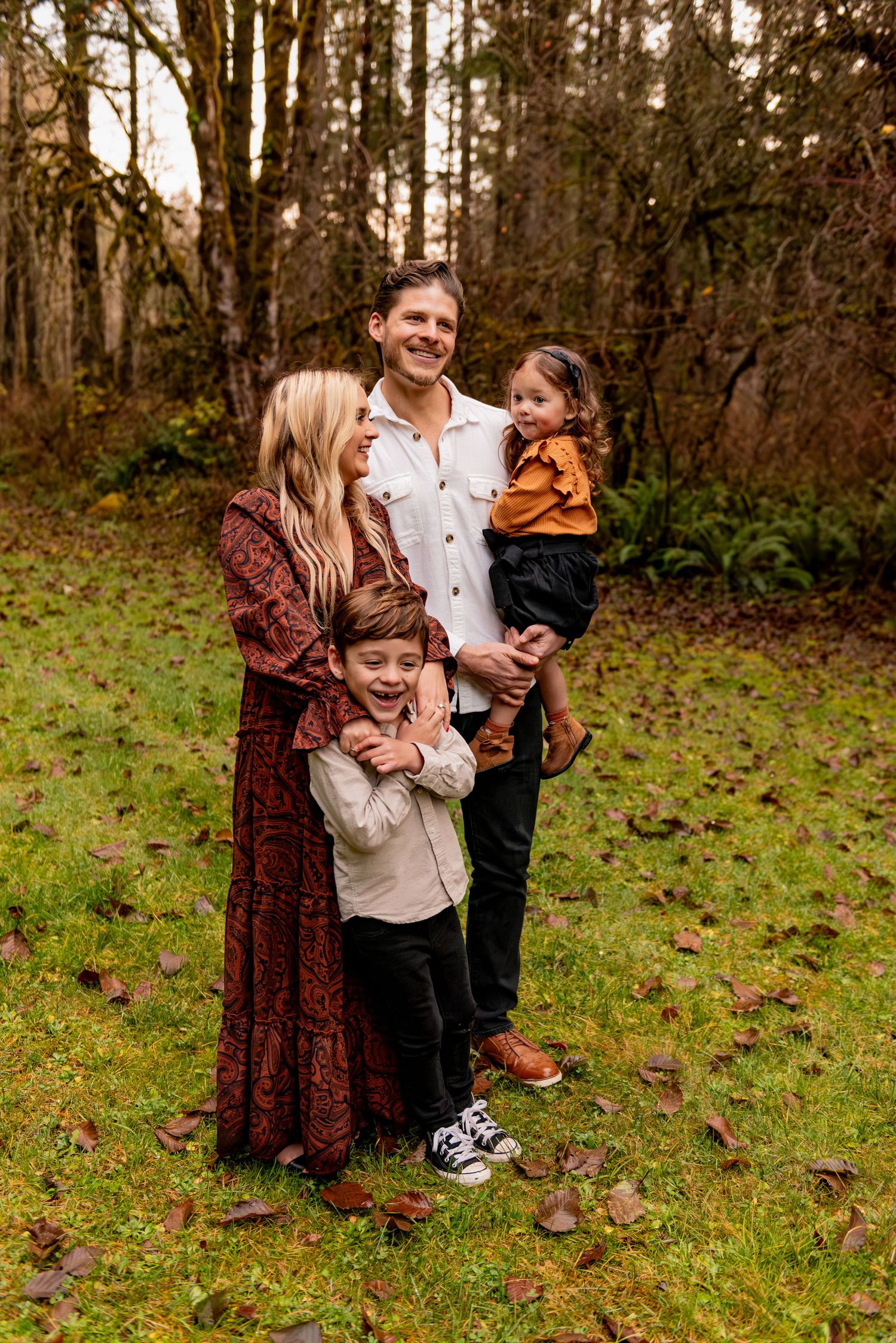 A family of four poses together outdoors in autumn, wearing coordinated casual outfits against a wooded backdrop. Silverton, OR