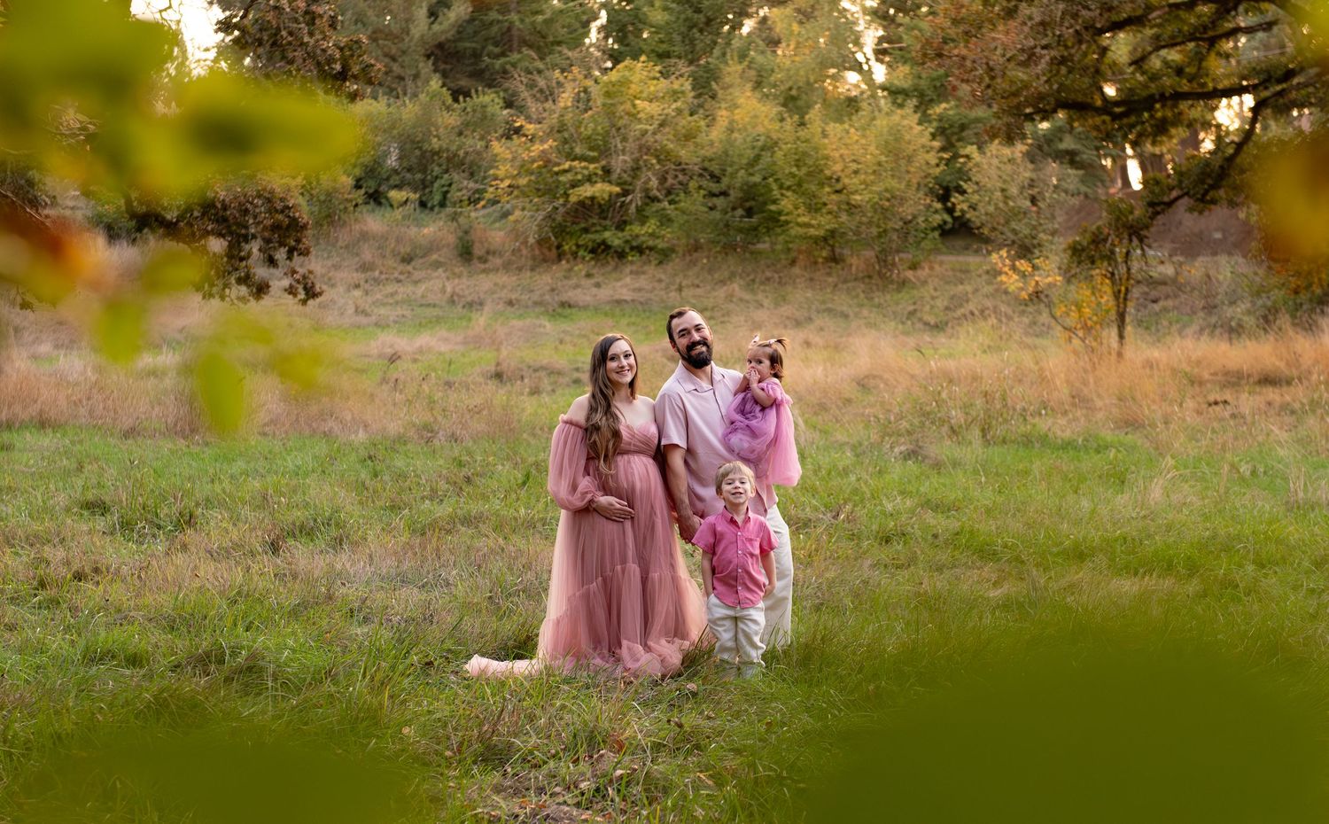 Family of four portrait located in a field with fall colored trees in the background and natural golden hour light. Located in Salem, OR