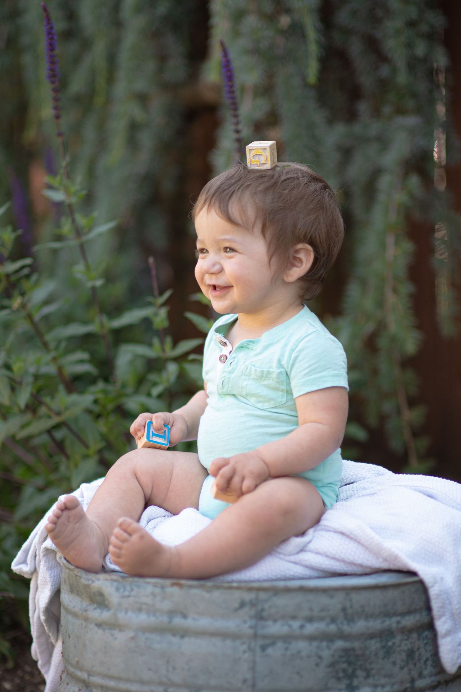 A toddler in a mint green shirt sits on a rustic metal tub in a garden setting with blooming flowers.