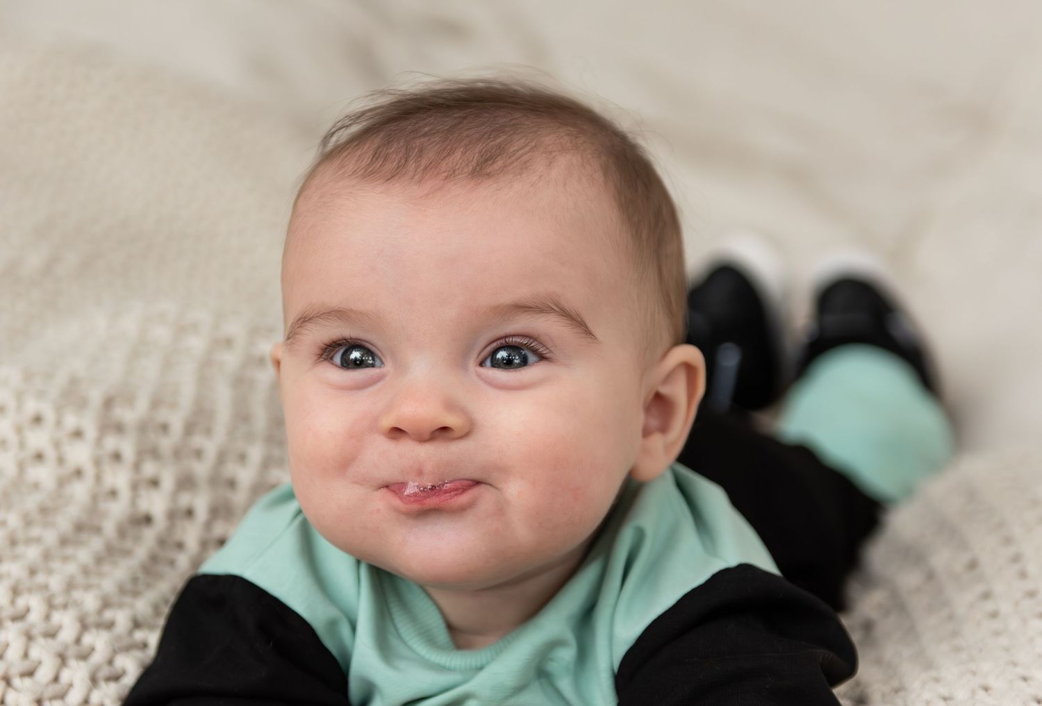 Six month milestone image with baby laying on a blanket, smiling and interacting with parents. Neutral backdrop with lettering. Salem, OR.