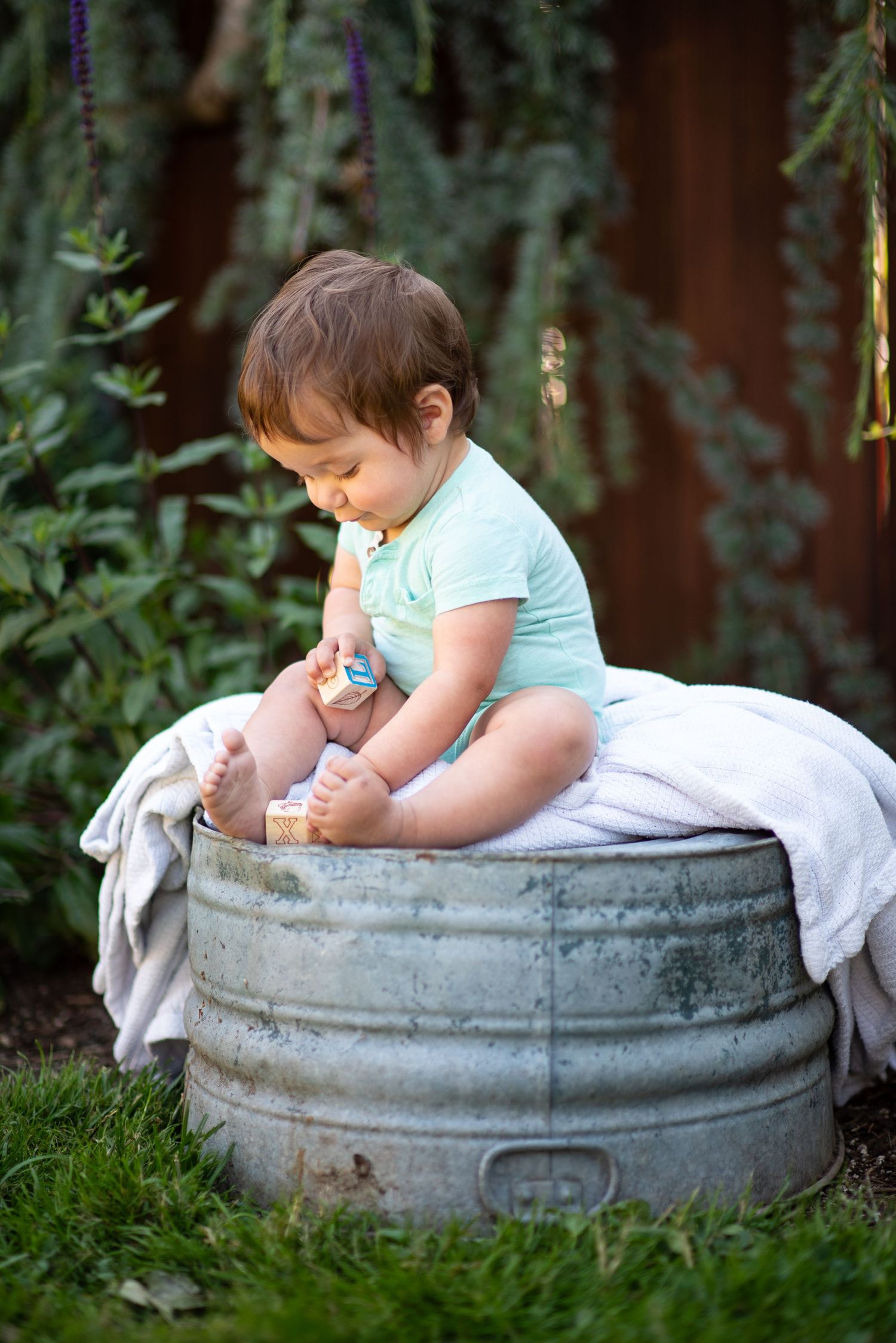 A small child in light clothing sits on a vintage galvanized wash tub in a garden setting.