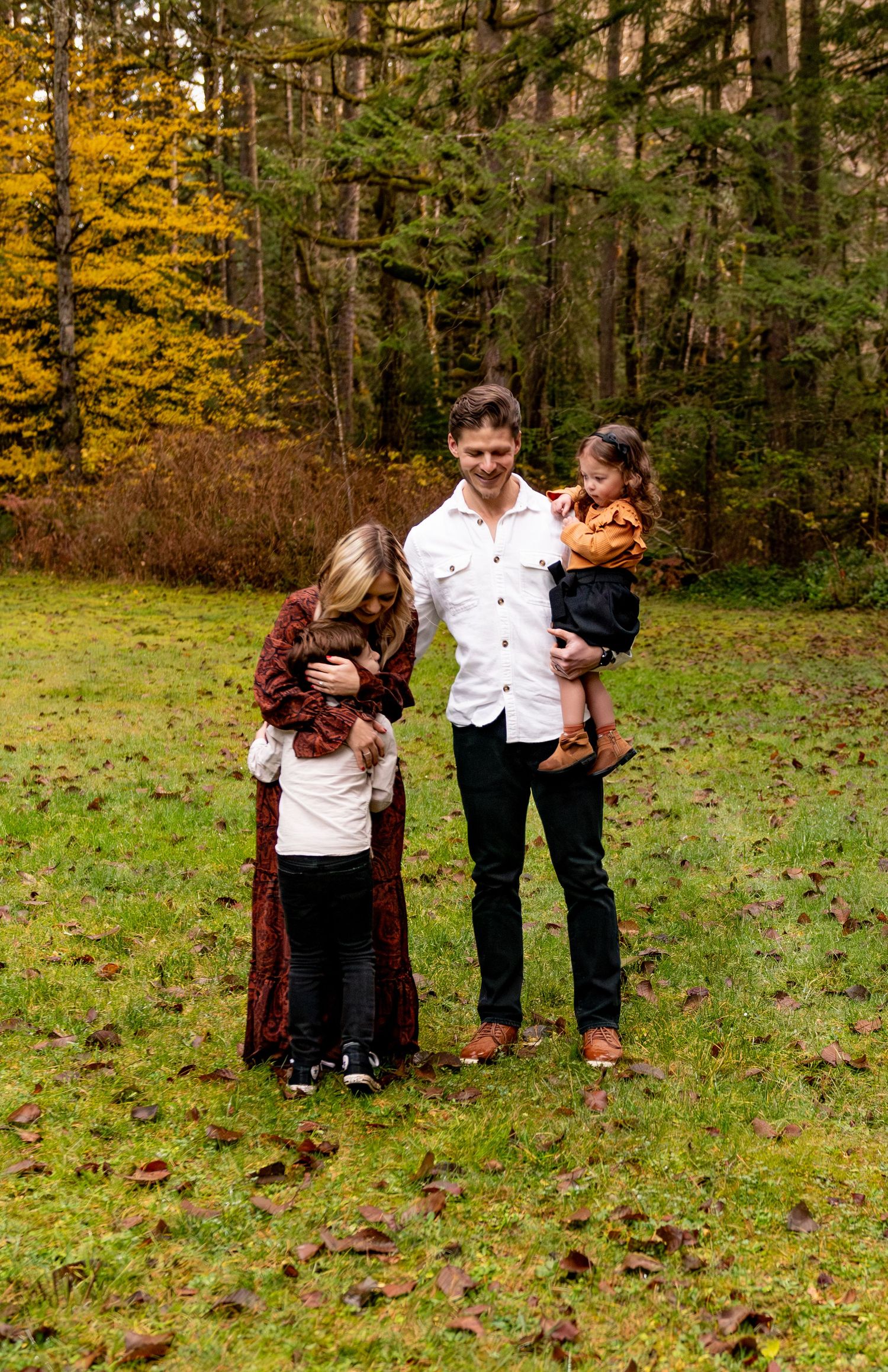 A family stands together on grass in an autumn forest setting with golden leaves and evergreen trees in the background. Silverton, OR