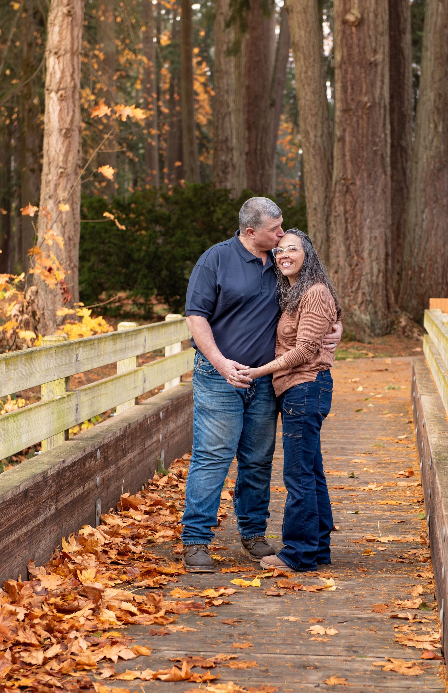 Two people share a tender moment while walking along a wooden bridge covered in autumn leaves in a forest setting. Stayton, OR