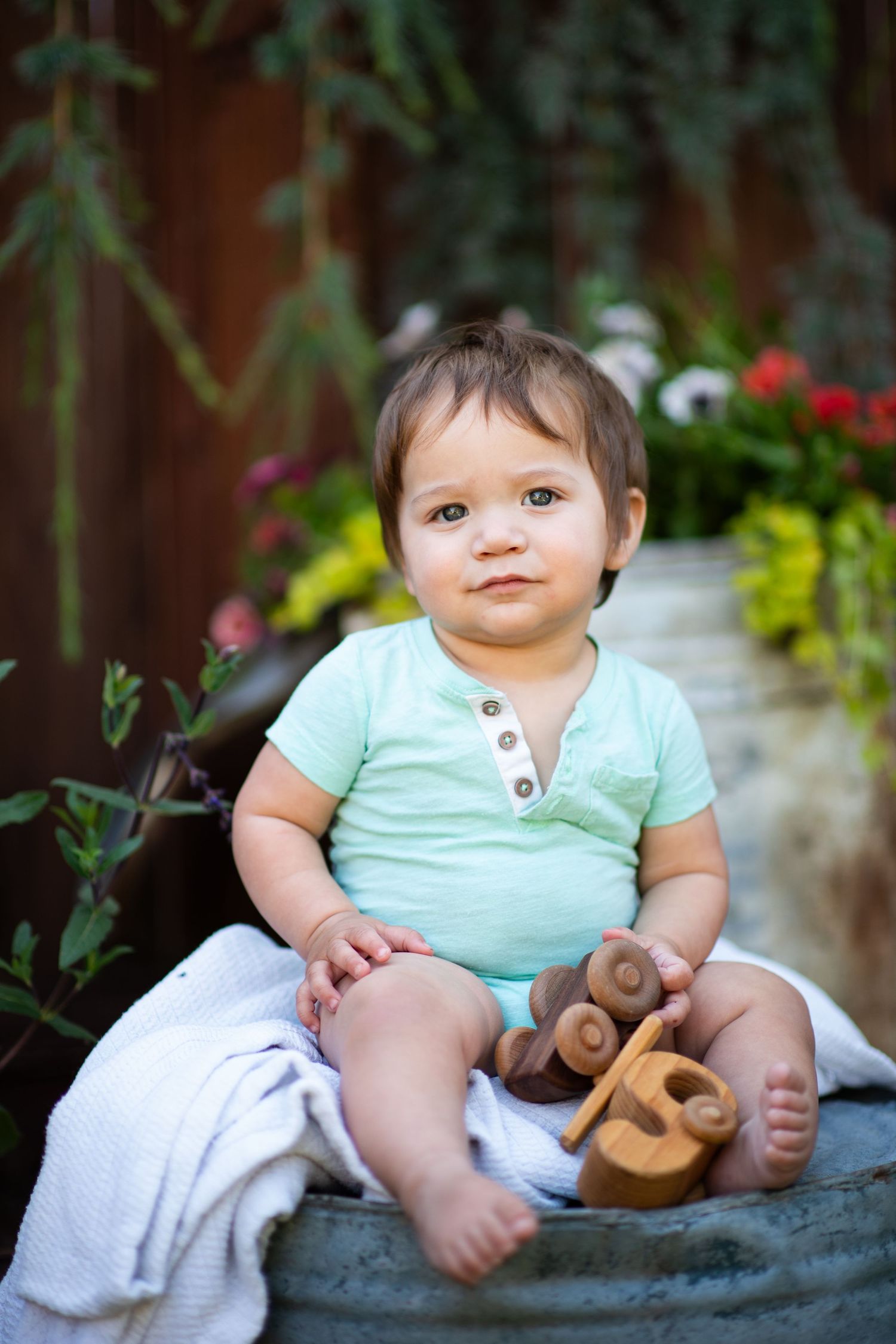 A small child in a mint green shirt sits on a blanket with a plush toy in a garden setting.