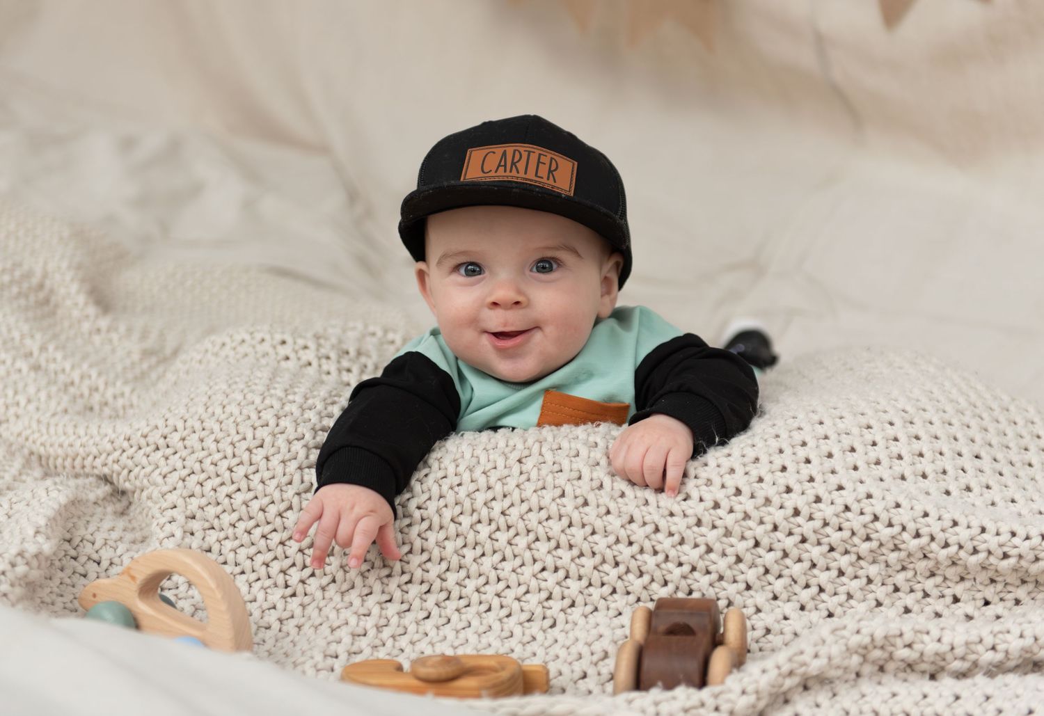 Six month milestone image with baby laying on a blanket, smiling and interacting with parents. Neutral backdrop with lettering. Salem, OR.
