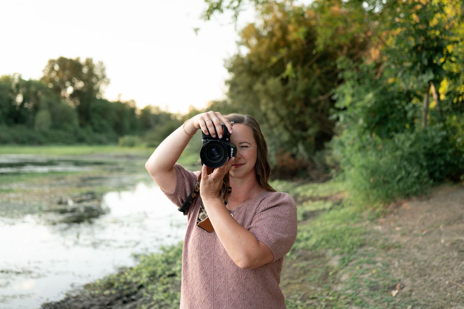 A photographer in a pink shirt takes photos near a serene pond at sunset with trees in the background. Salem, OR