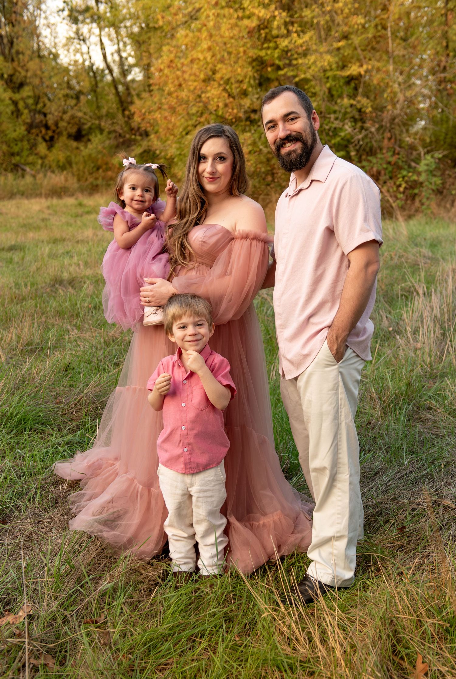 Family of four portrait located in a field with fall colored trees in the background and natural golden hour light. Located in Salem, OR