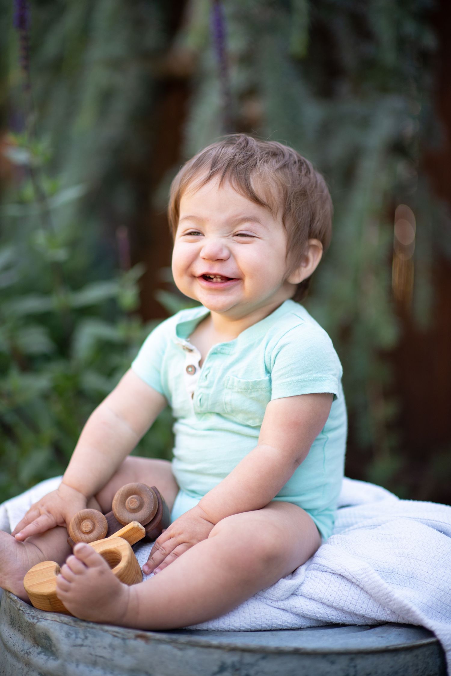 A baby in a light blue outfit lies on a white blanket while playing with wooden toys and laughing joyfully.