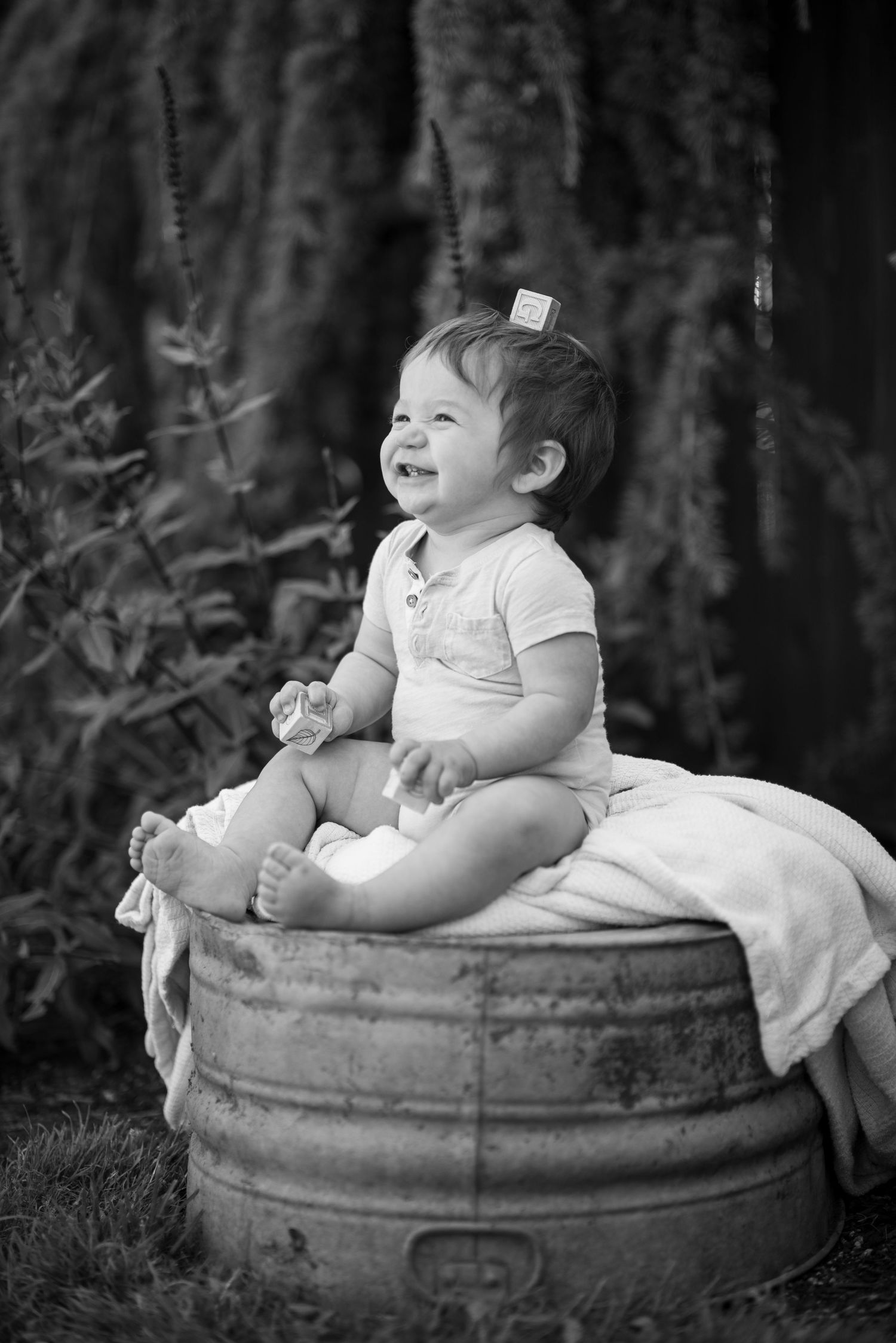 Black and white photo series of a baby in white clothing sitting and laughing on a rustic barrel outdoors.