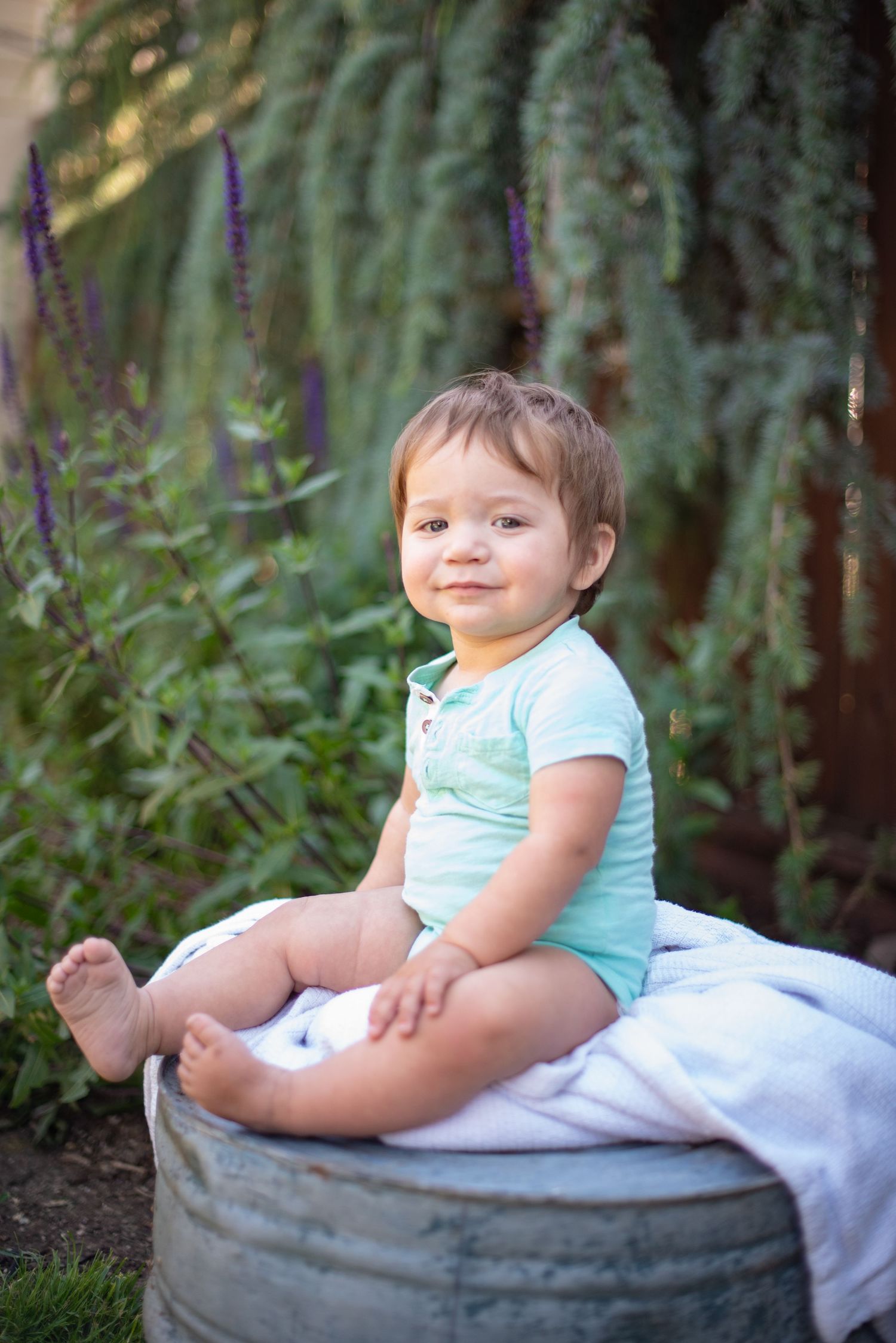 A baby  sits on a large basin outdoors with greenery in the background.