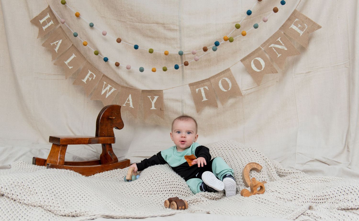 Six month milestone image with baby laying on a blanket, smiling and interacting with parents. Neutral backdrop with lettering. Salem, OR.