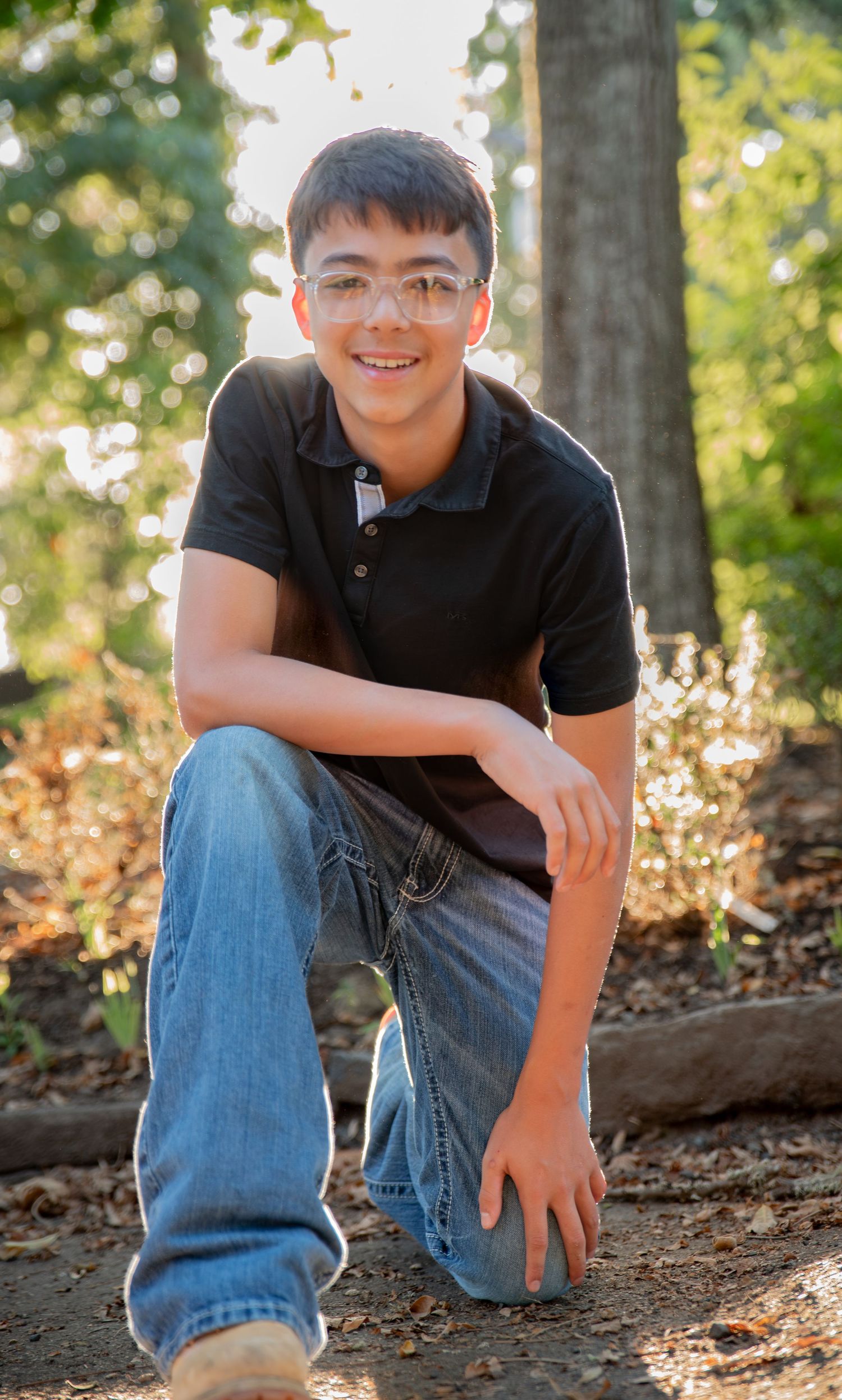Young teen boy, wearing glasses,  kneeling with a backlit sun among trees in a park located in Salem, OR.