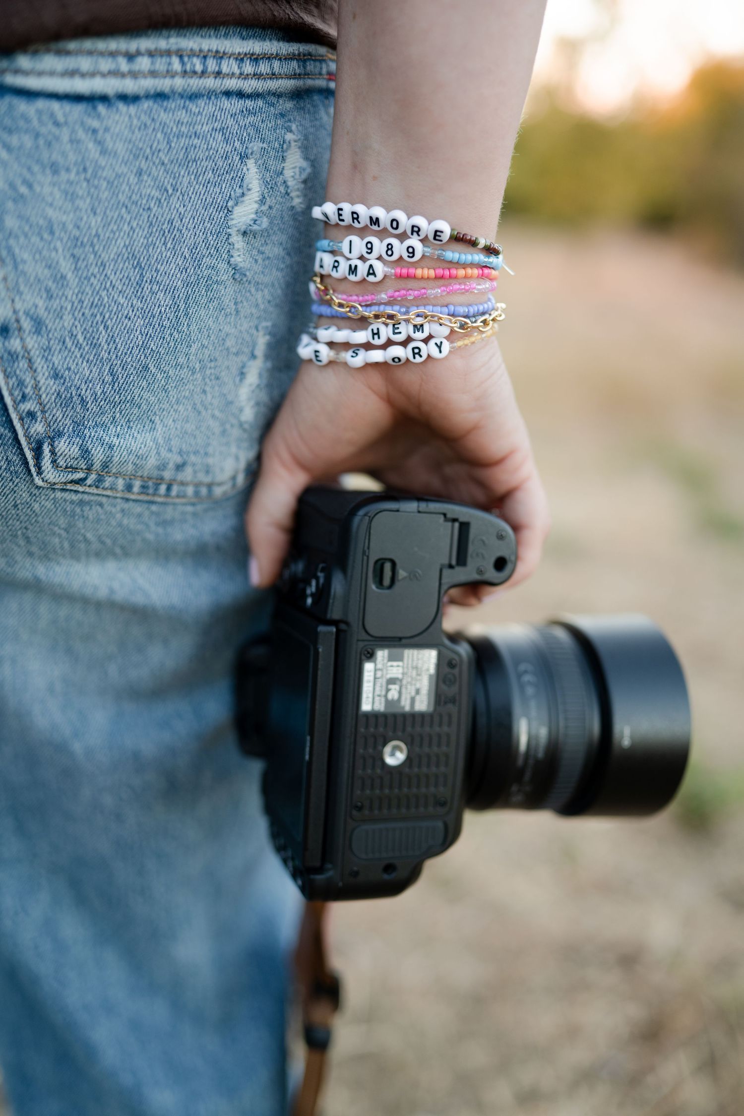 Hand wearing bracelets holds a black DSLR camera with telephoto lens against denim background.