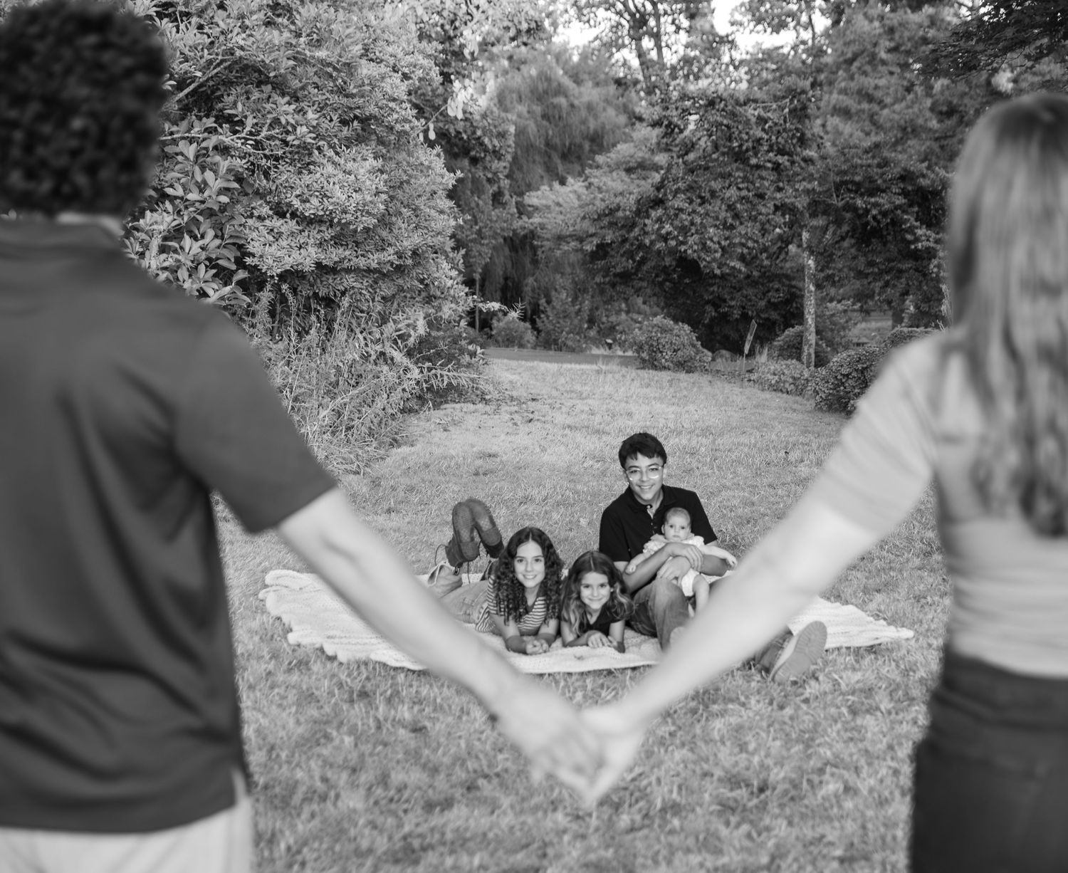 Mom and dad holding hands in a black and white photo with the kids laying on a blanket in the background surrounded by trees. Salem, OR.