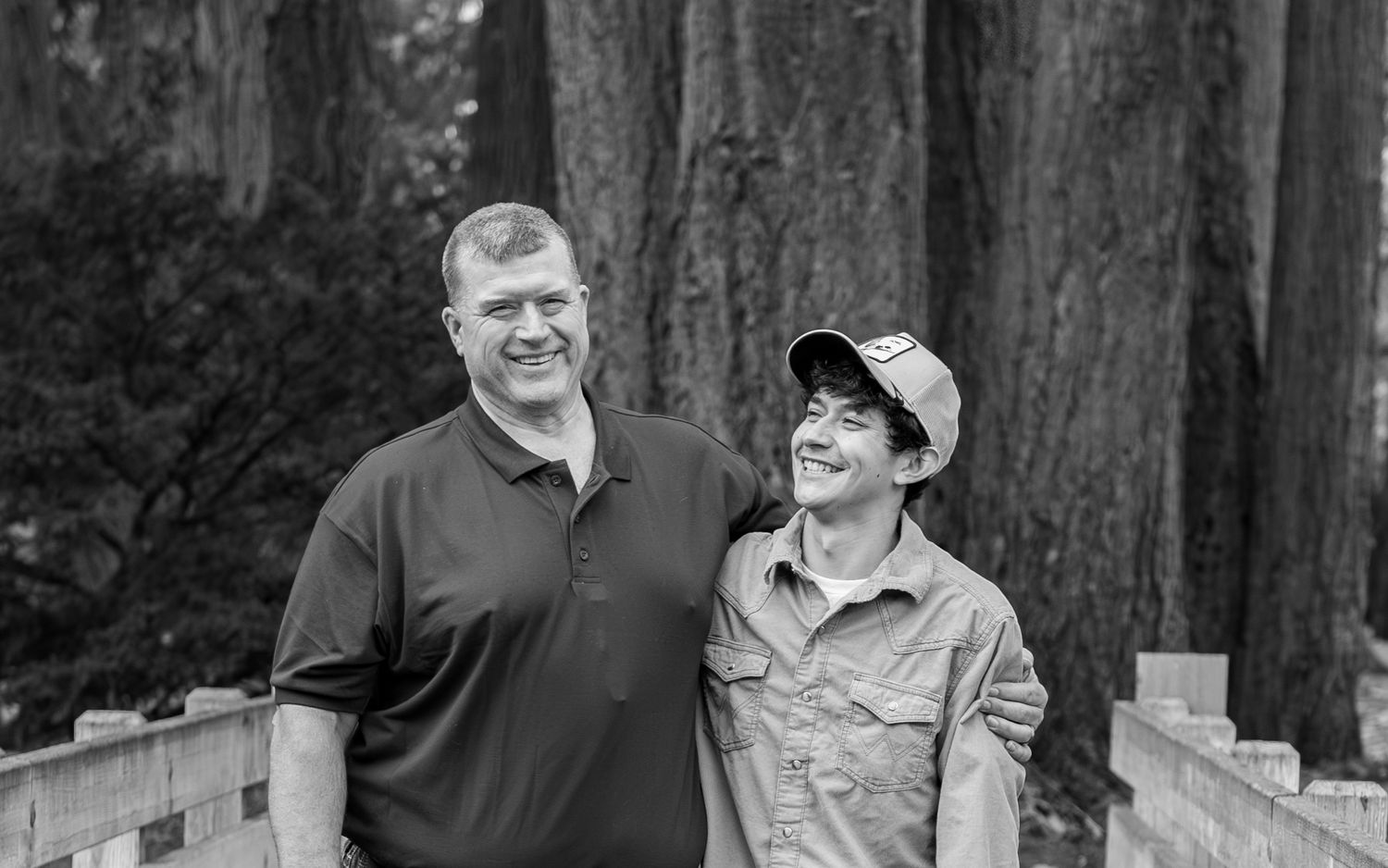 Two people stand together smiling on a wooden walkway surrounded by tall redwood trees in a black and white photo. Stayton, OR