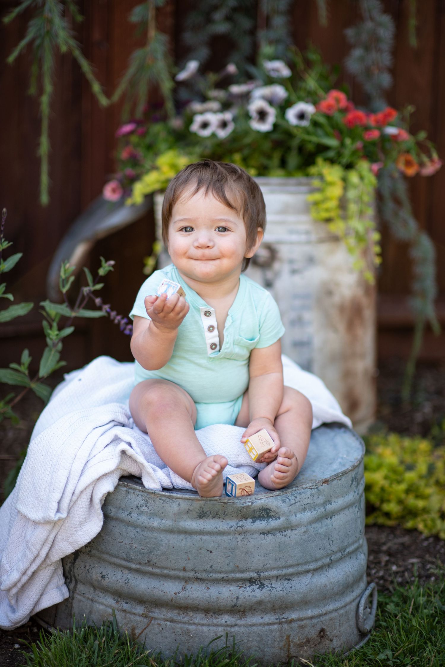 Small child sits in a rustic concrete planter surrounded by garden greenery and flowers in outdoor setting.