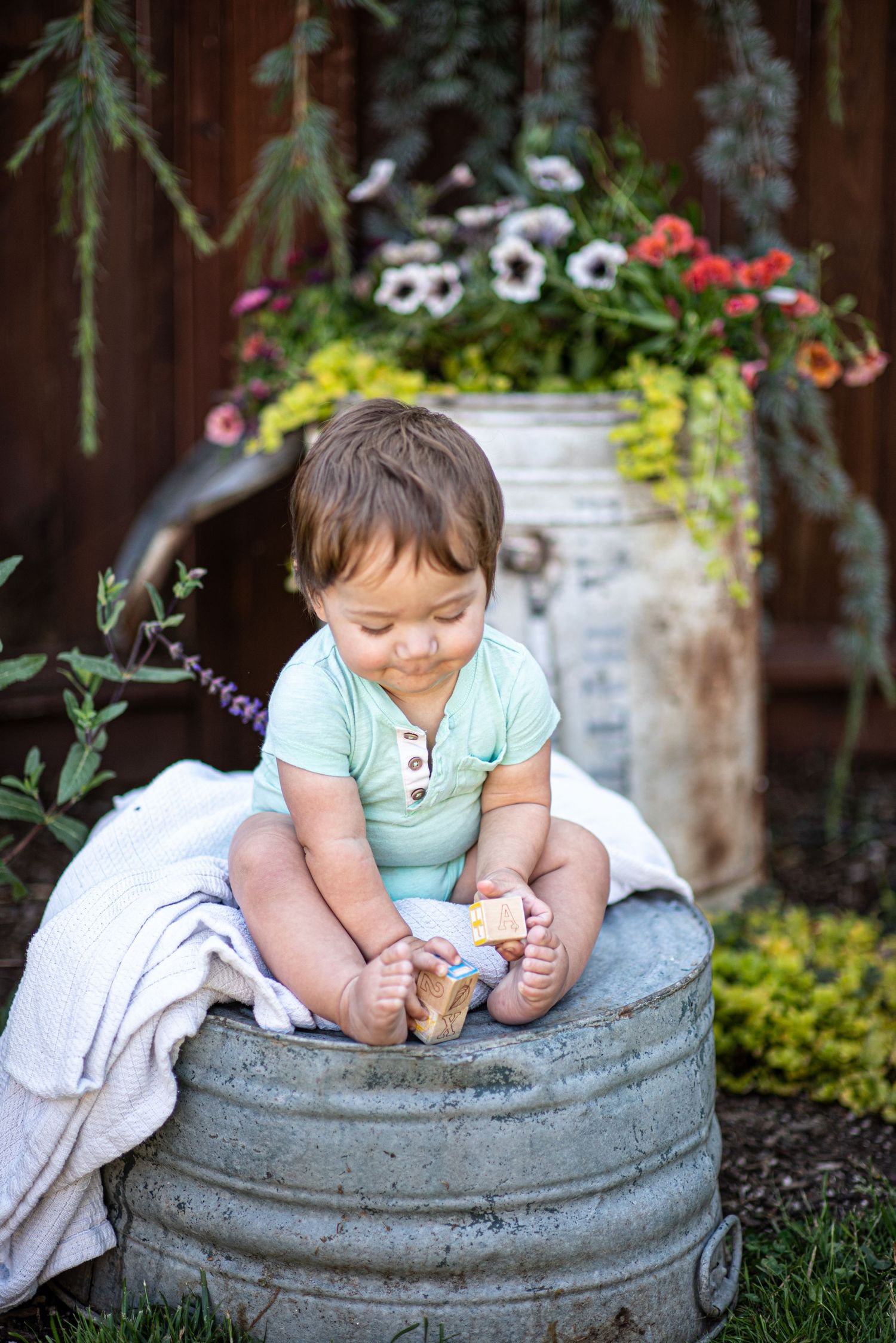 A toddler in a mint green shirt sits on a rustic metal tub in a garden setting with blooming flowers.