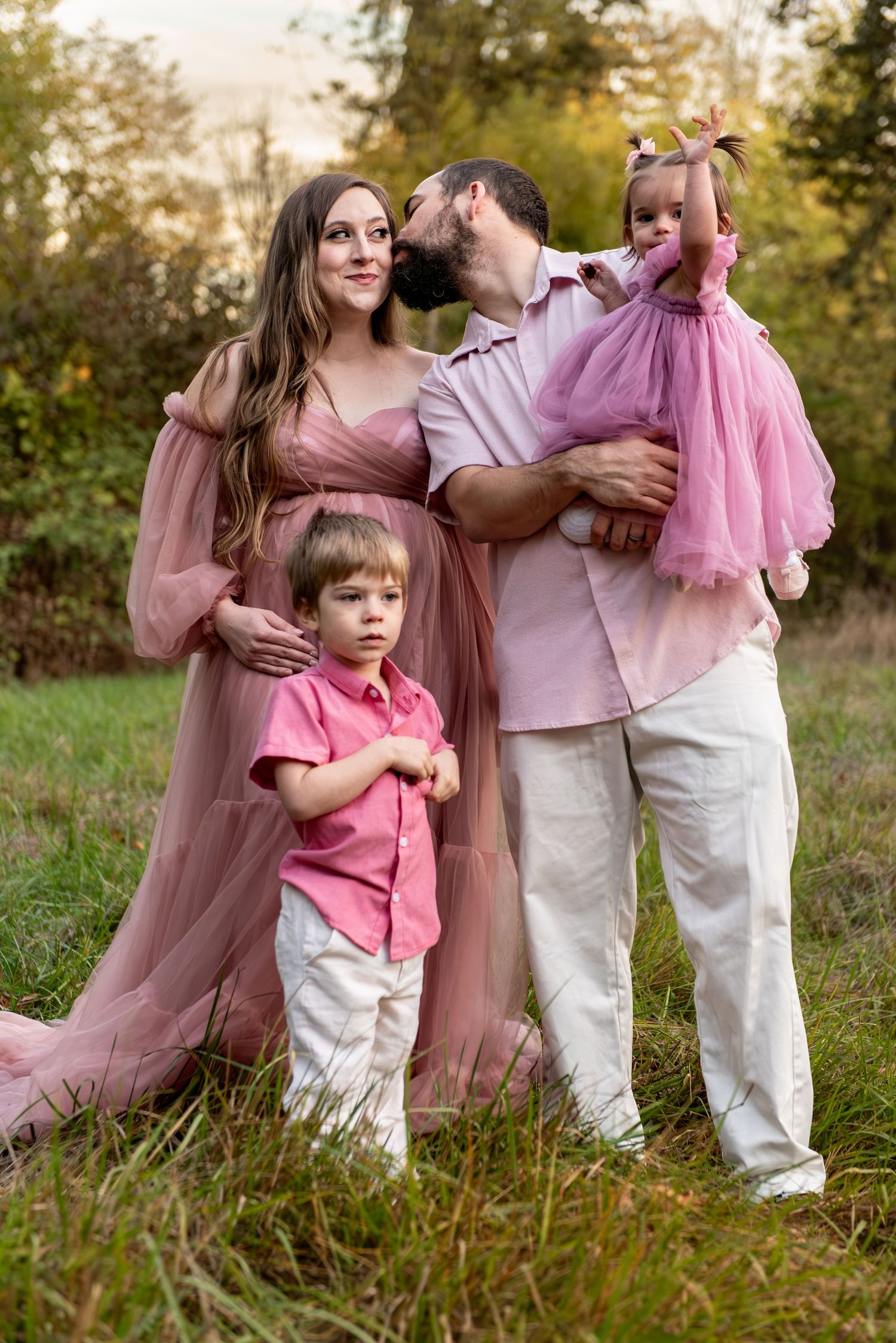 Dad kissing Mom holding daughter and son is standing on the ground in a family image in a field in Salem, OR