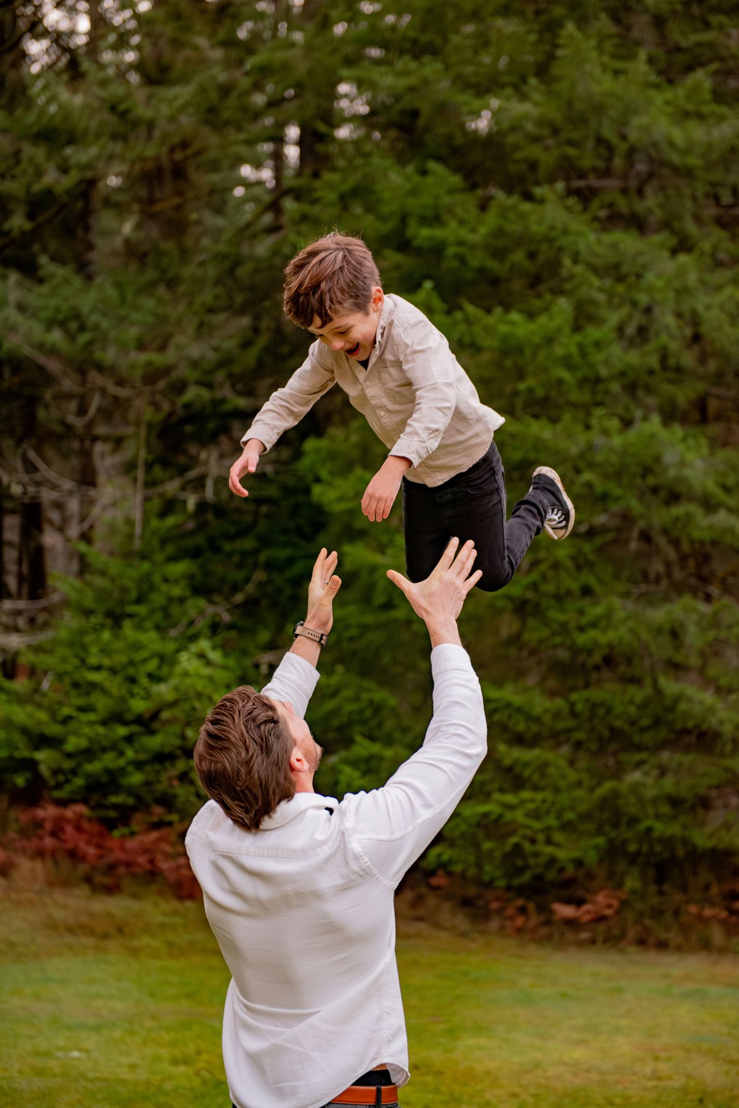 A person in a white shirt lifts and tosses a small child playfully in the air on a grassy lawn with trees in background. Silverton, OR