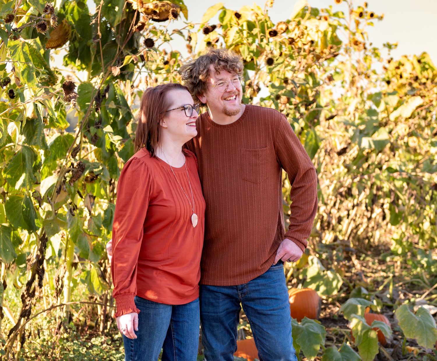 A couple in coordinating rust-colored sweaters laughs together while walking through an autumn pumpkin patch.