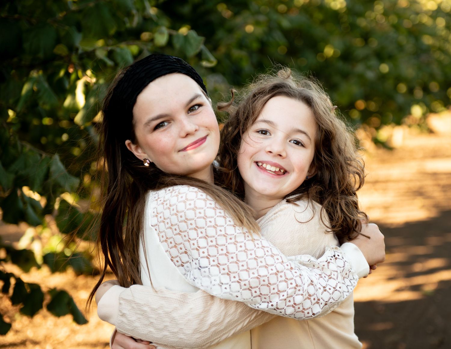 Two people embracing and smiling warmly on a sunlit outdoor path with green foliage in the background.