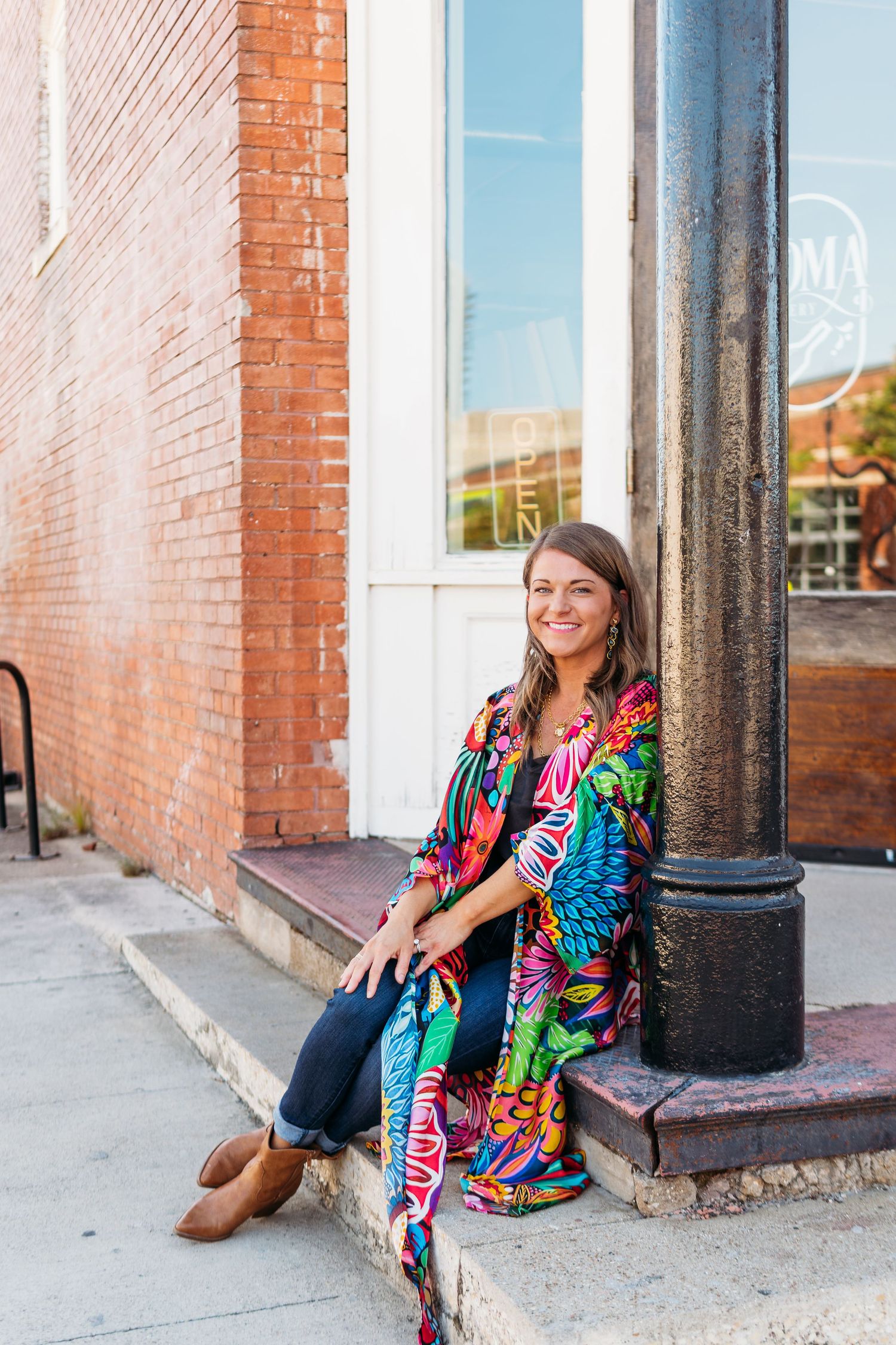 A woman sitting outdoors wearing a long kimono with a vibrant print displaying a clothing item for a brand.
