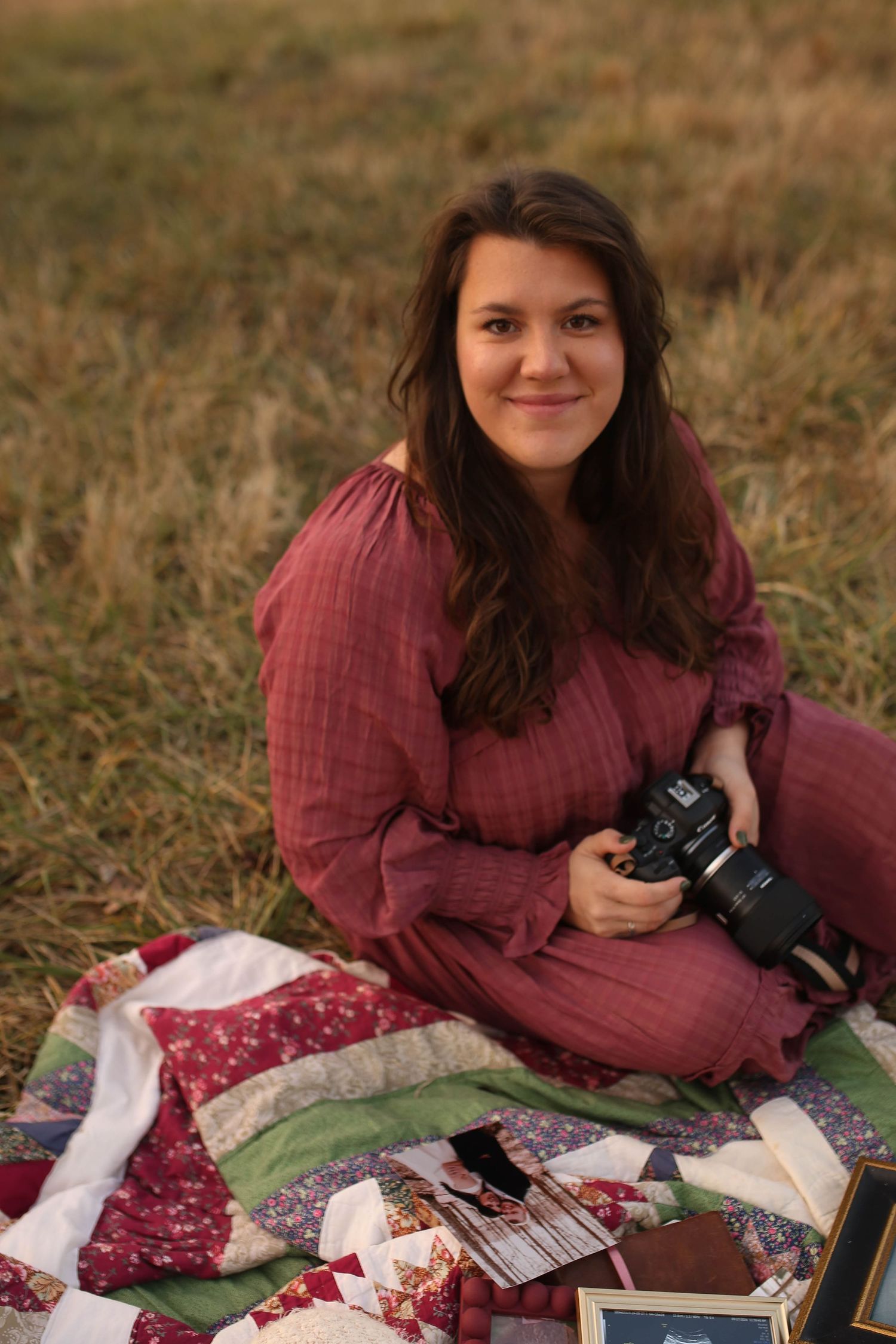 Young woman in burgundy dress sitting on a patchwork quilt in a field, holding a camera, smiling softly.