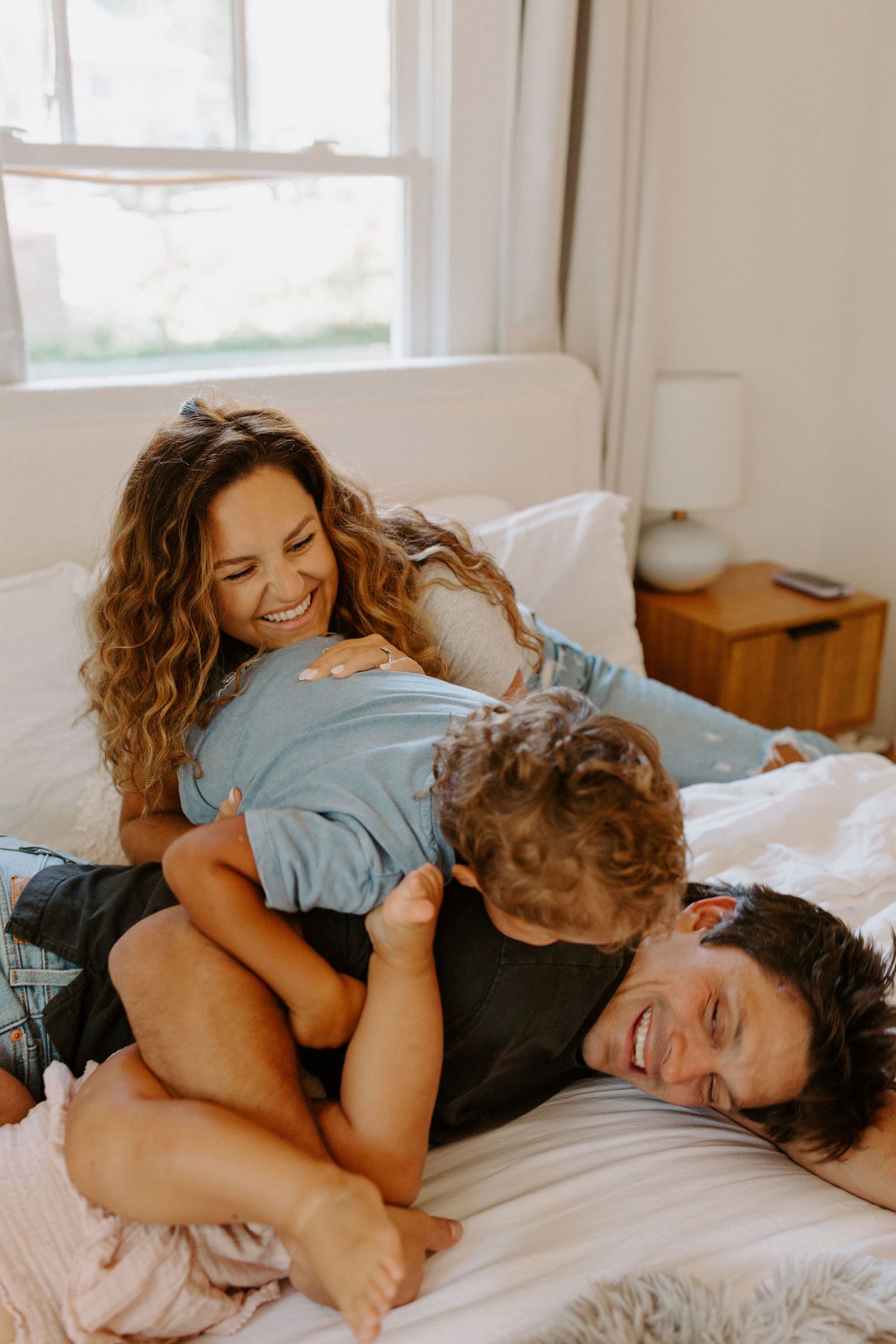 Intimate family playtime captured on a sunlit bed with white linens.