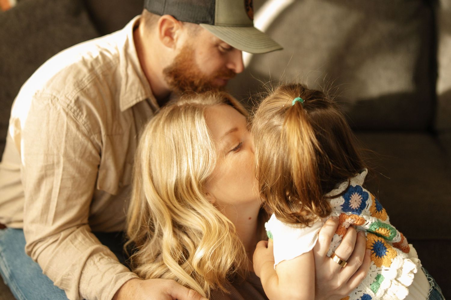 A loving family moment as parents kiss their young daughter on the cheeks in a warm, sunlit setting.