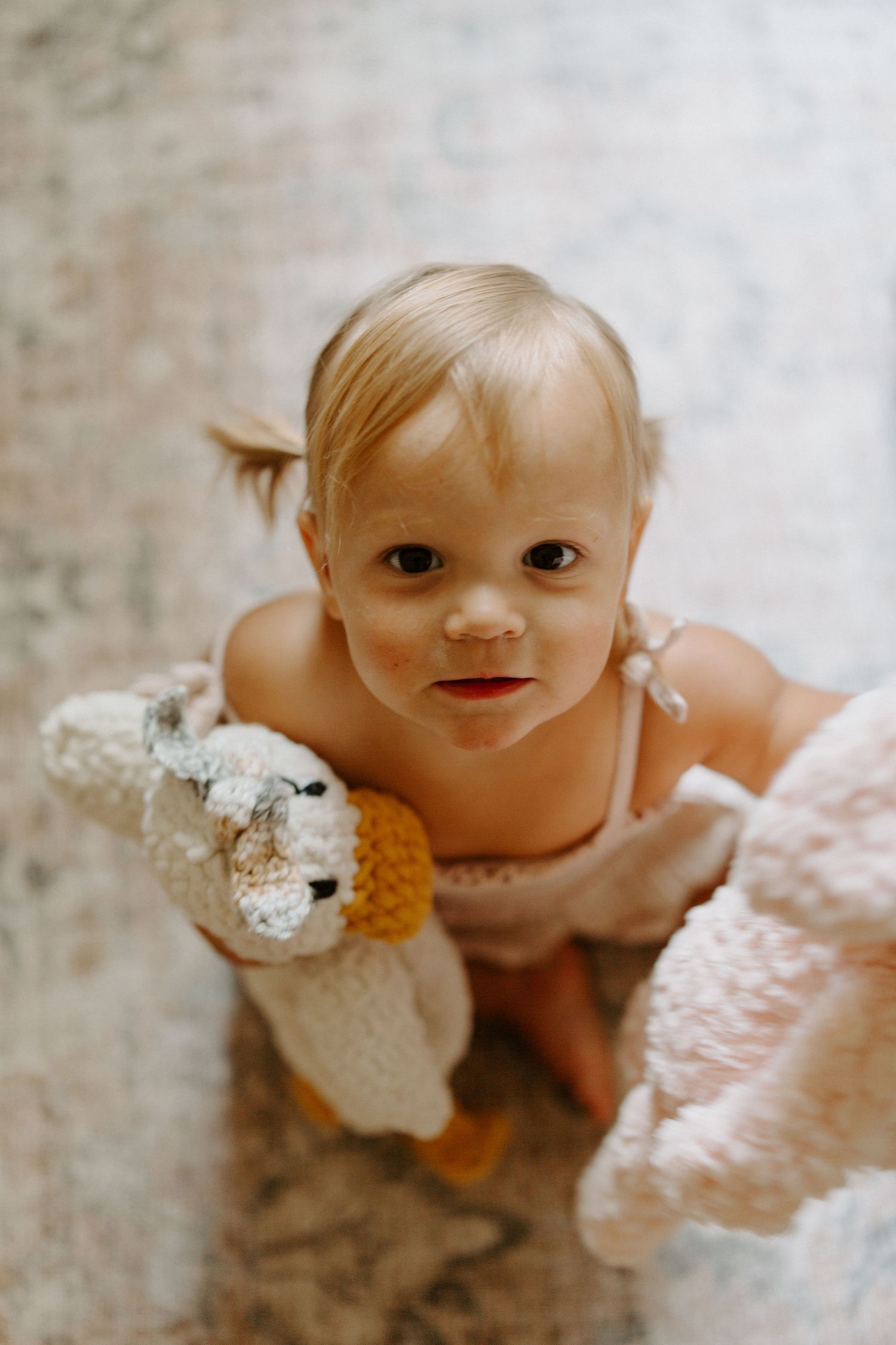 A toddler in a peach outfit holds a stuffed animal while sitting on a patterned rug.