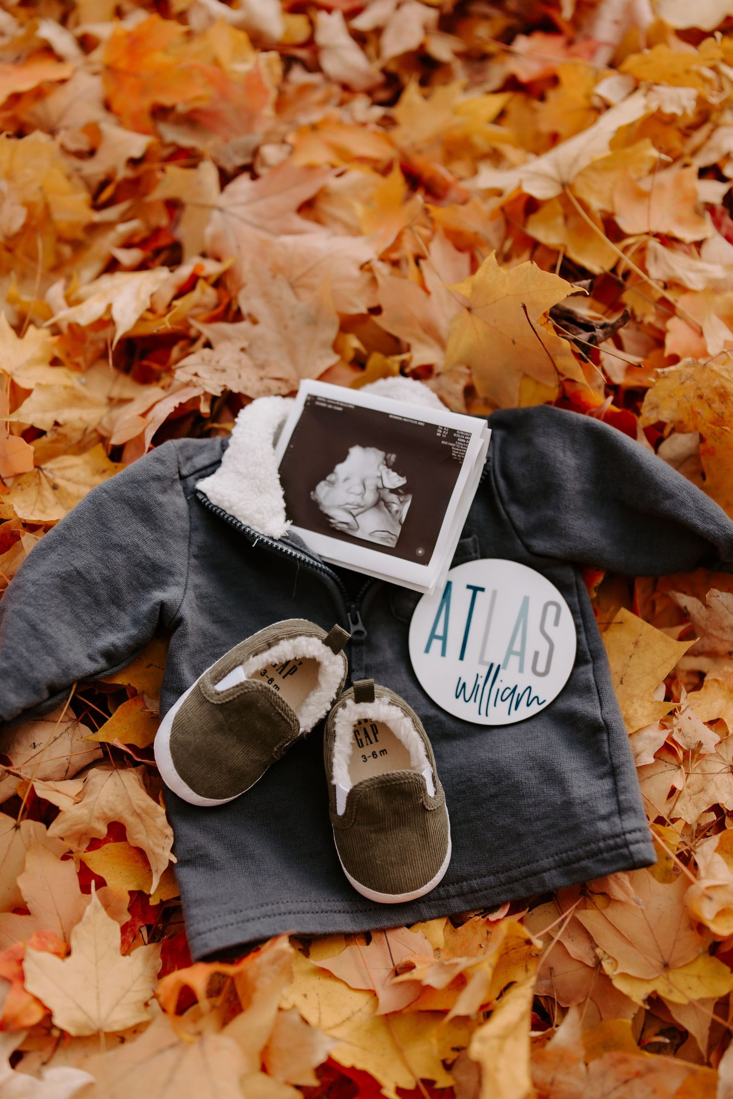 Baby shoes and ultrasound photos arranged on fallen autumn leaves with name 'Atlas' visible.