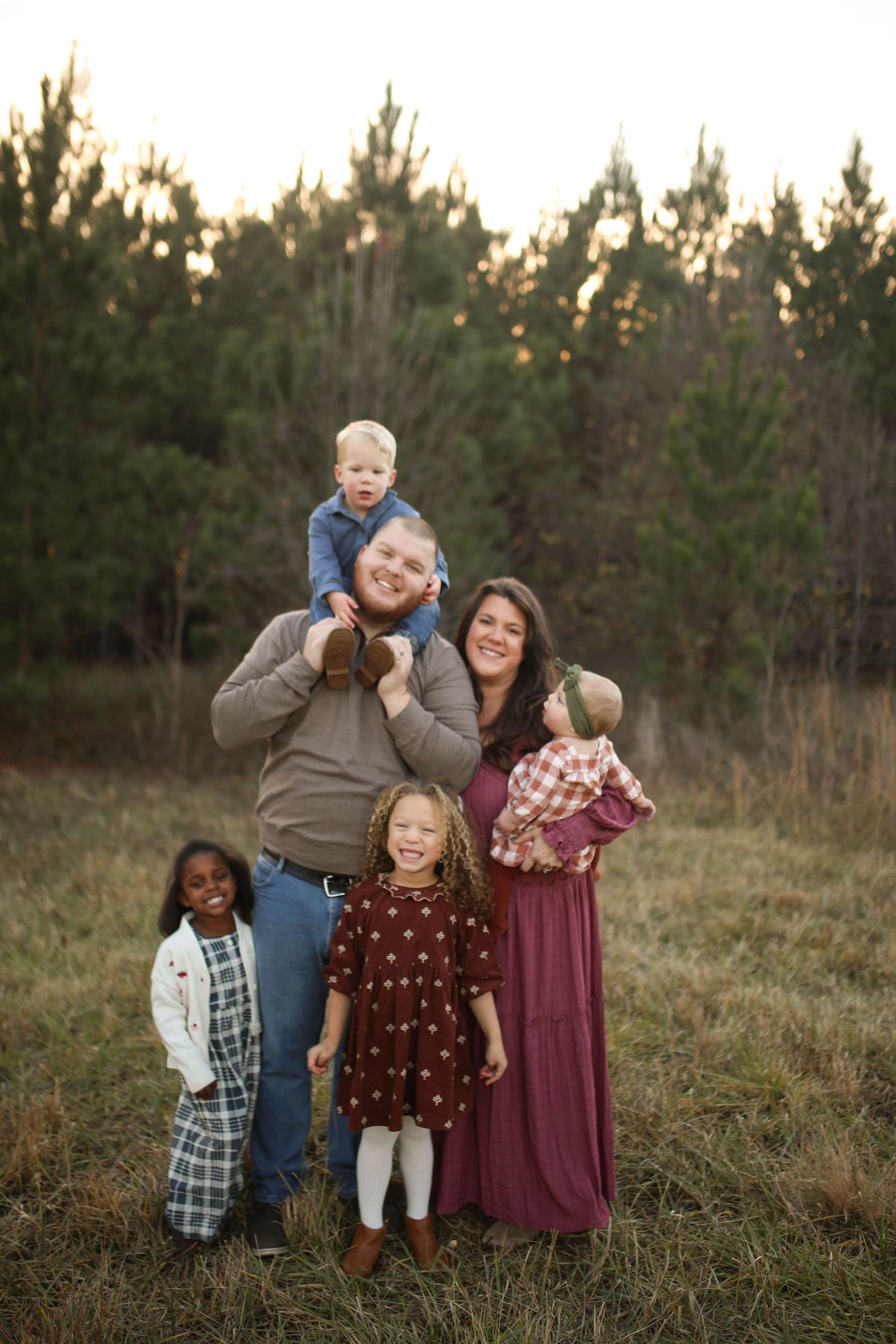 A joyful family of five poses outdoors in a field with pine trees, parents holding children in warm autumn attire.
