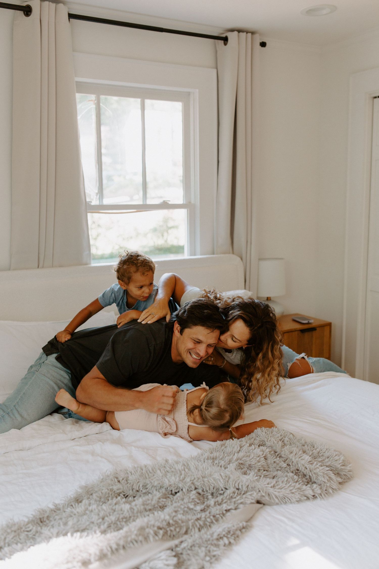 Intimate family cuddle session on a bed with soft natural lighting from the window.