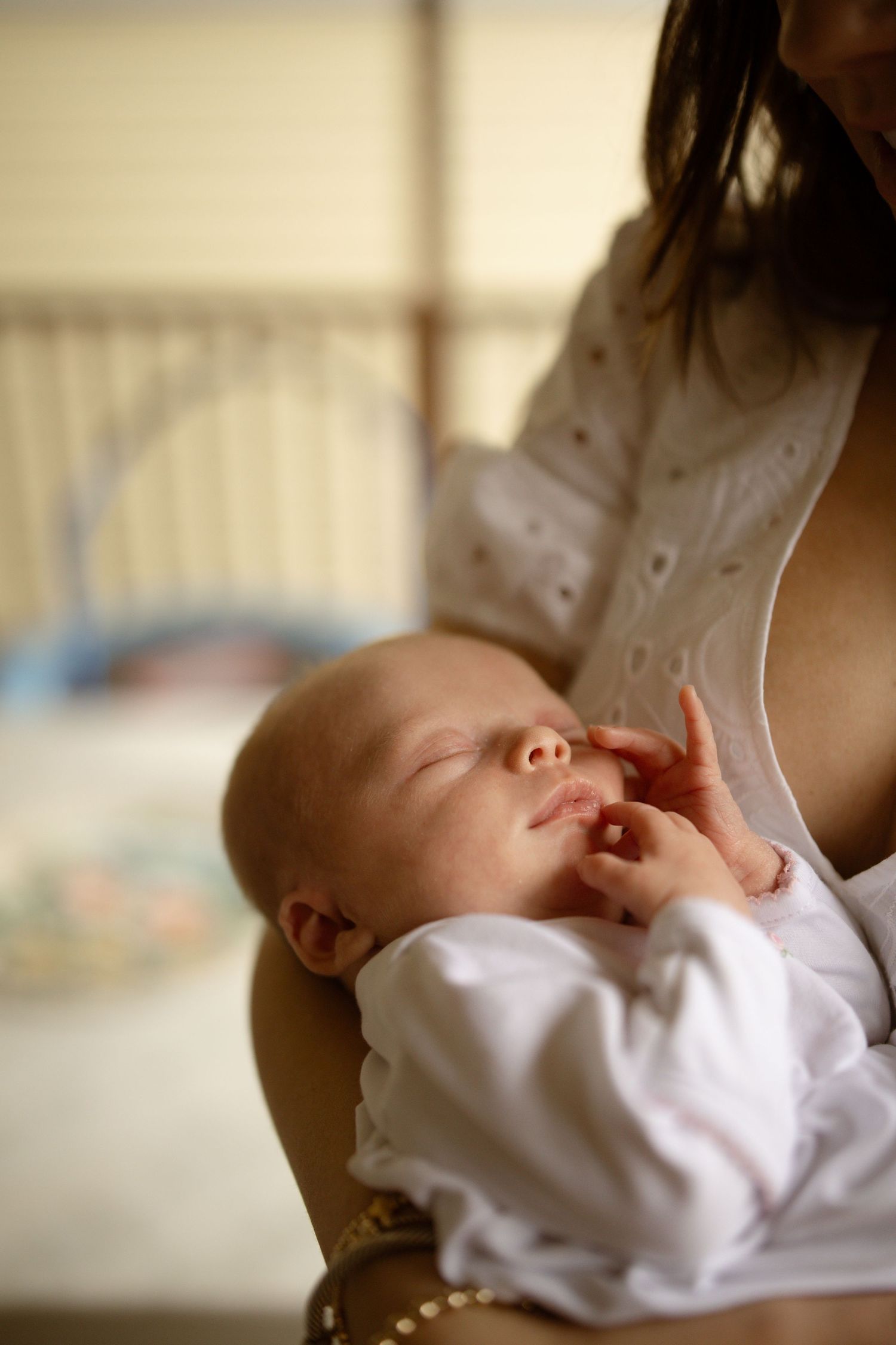 A peaceful newborn baby sleeping in mother's arms, dressed in white, with tiny hands raised near face.