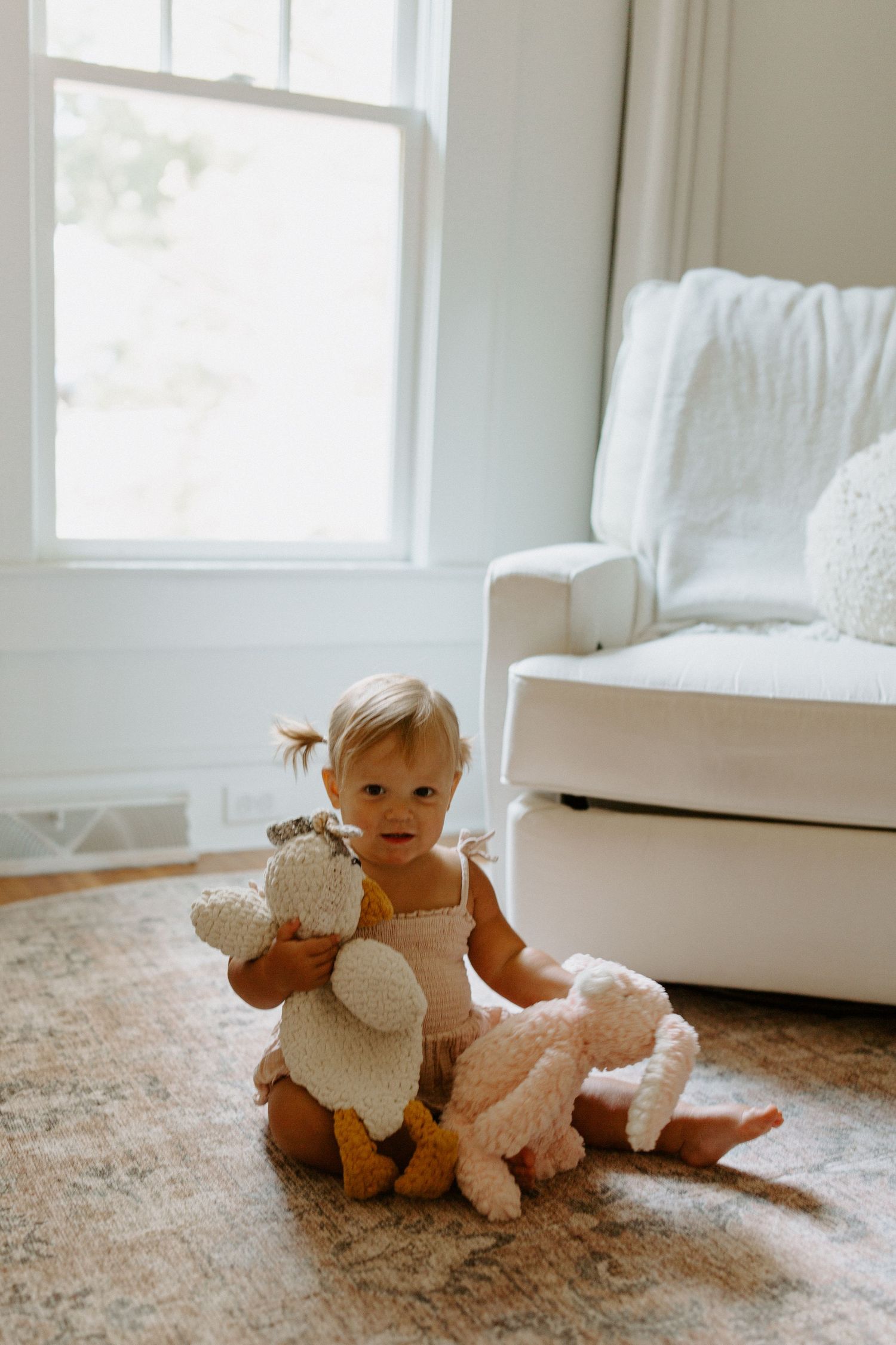 A toddler sits on a beige carpet playing with plush toys next to a white armchair by a window.