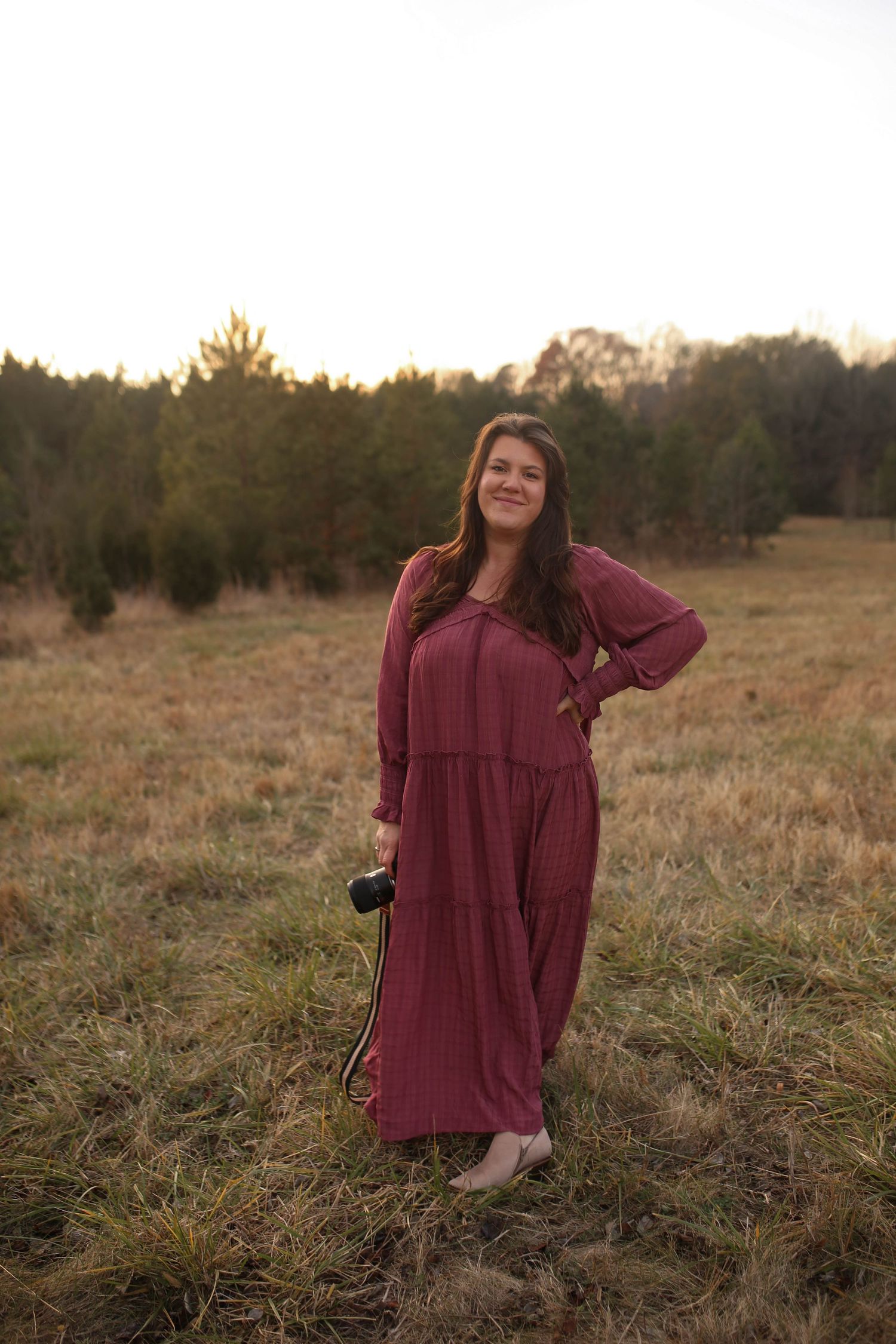 Woman in burgundy maxi dress holding camera standing in golden field at sunset with evergreen trees in background.