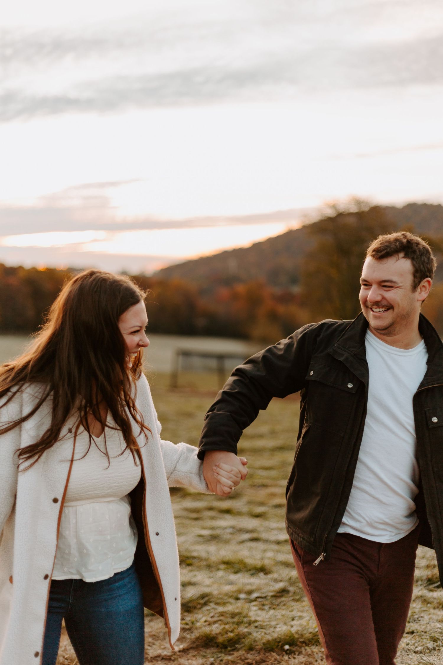 A playful couple laughs together in a field during golden hour with mountains in the background.