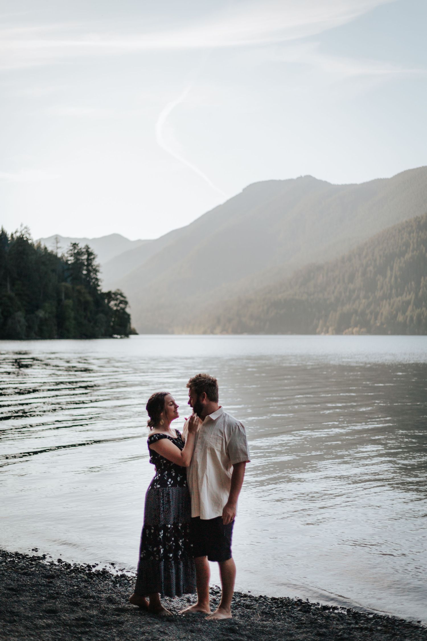 Couple sits together on rocky beach shore with mountain lake backdrop.