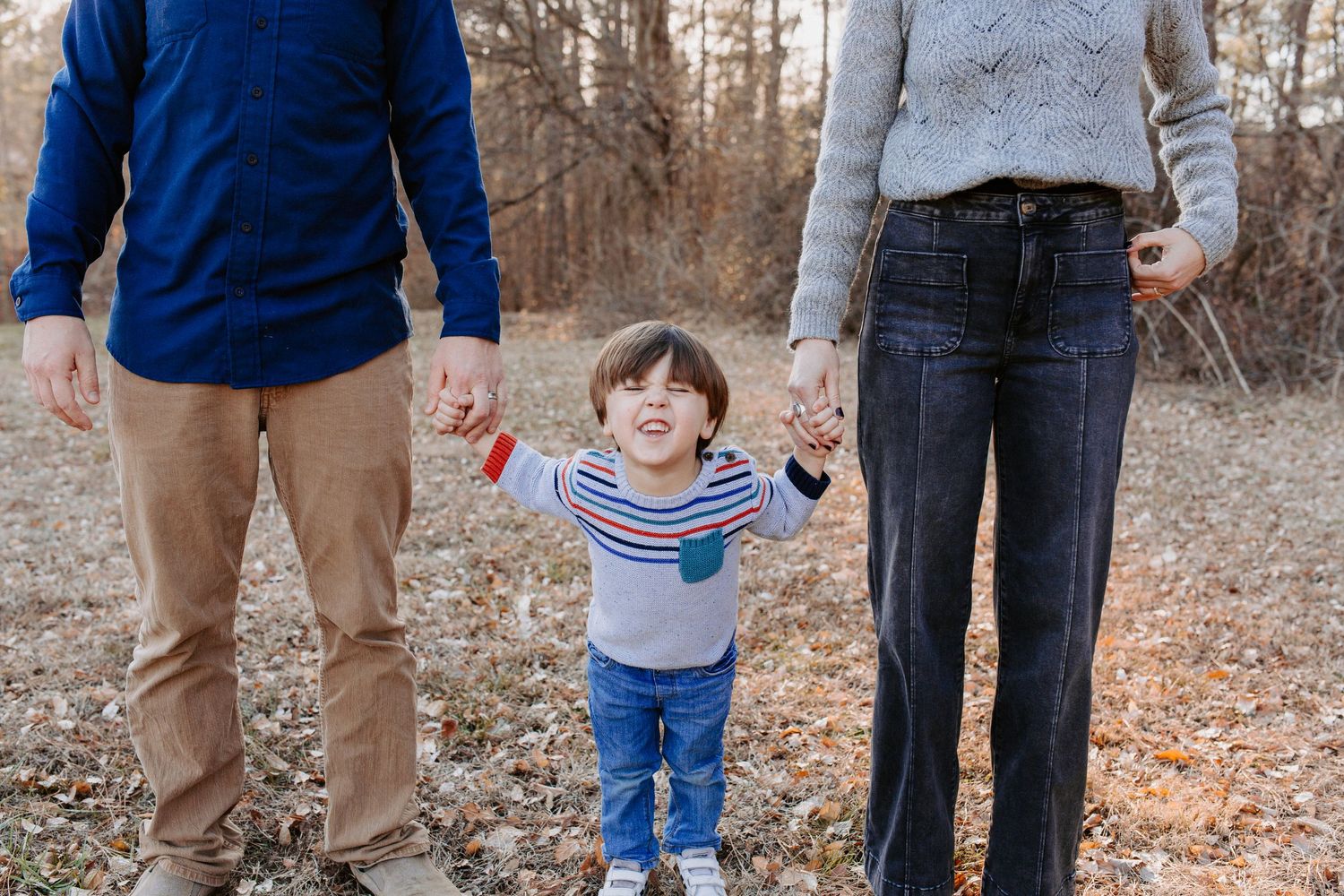 Child holding hands with parents while walking through autumn leaves in the woods.