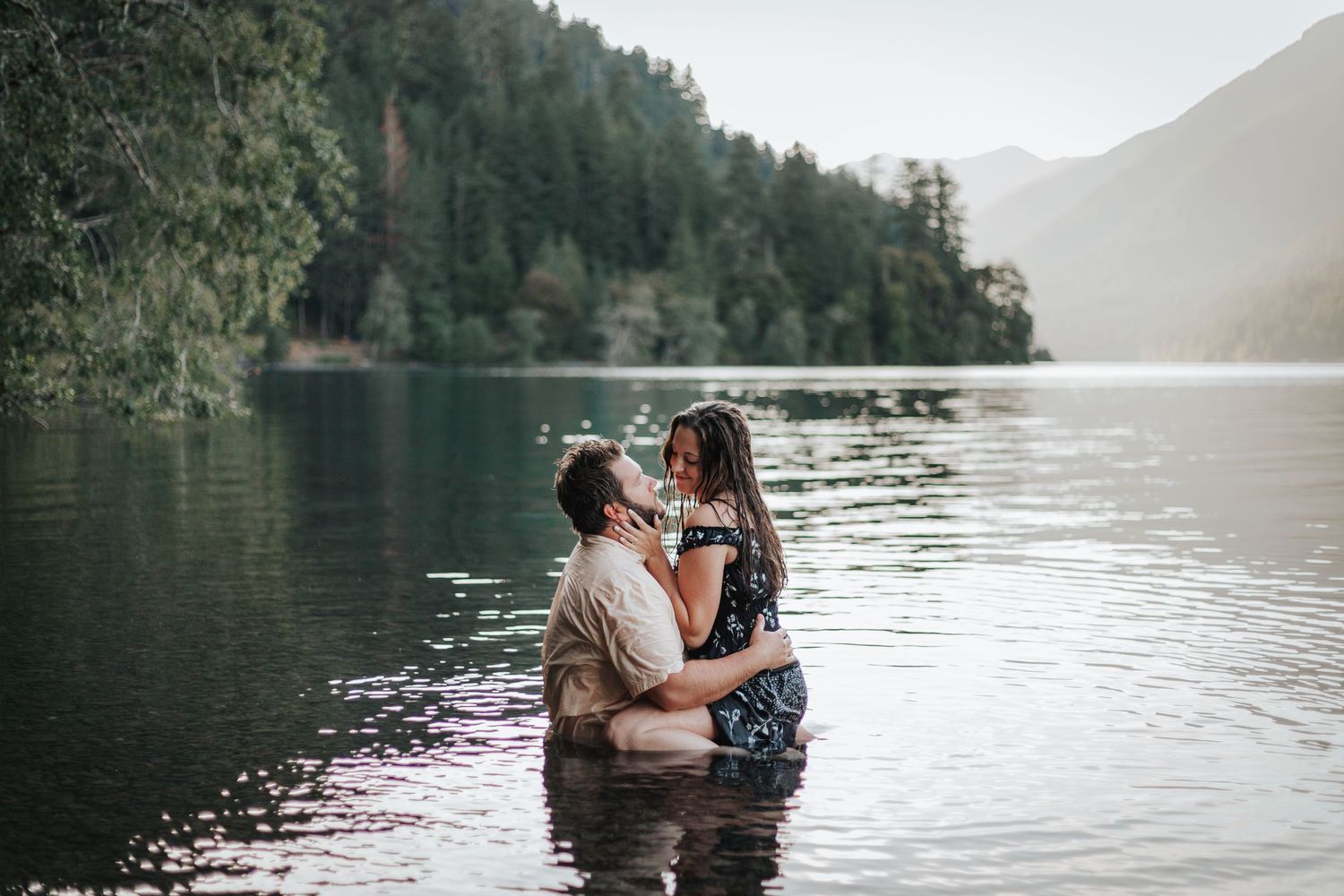 Two people share an intimate moment in a serene lake surrounded by evergreen forests.