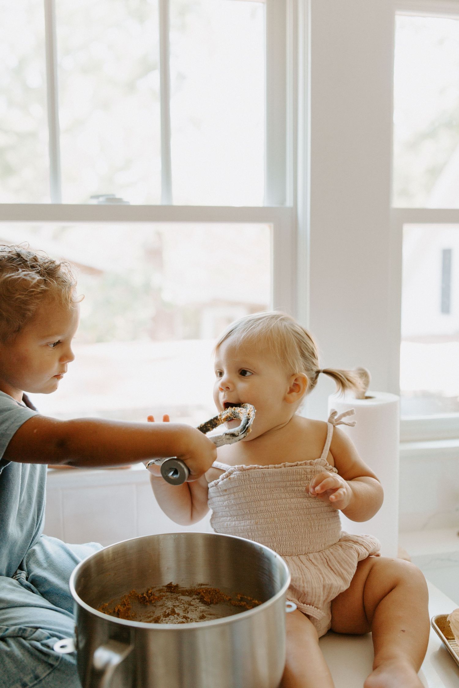 A toddler in a knit romper being fed from a mixing bowl in a sunny kitchen.