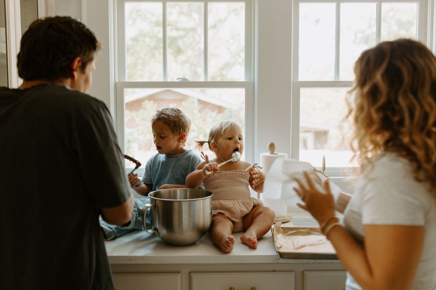 Family cooking together in bright sunlit kitchen with children sitting on countertop while mixing ingredients in bowl.