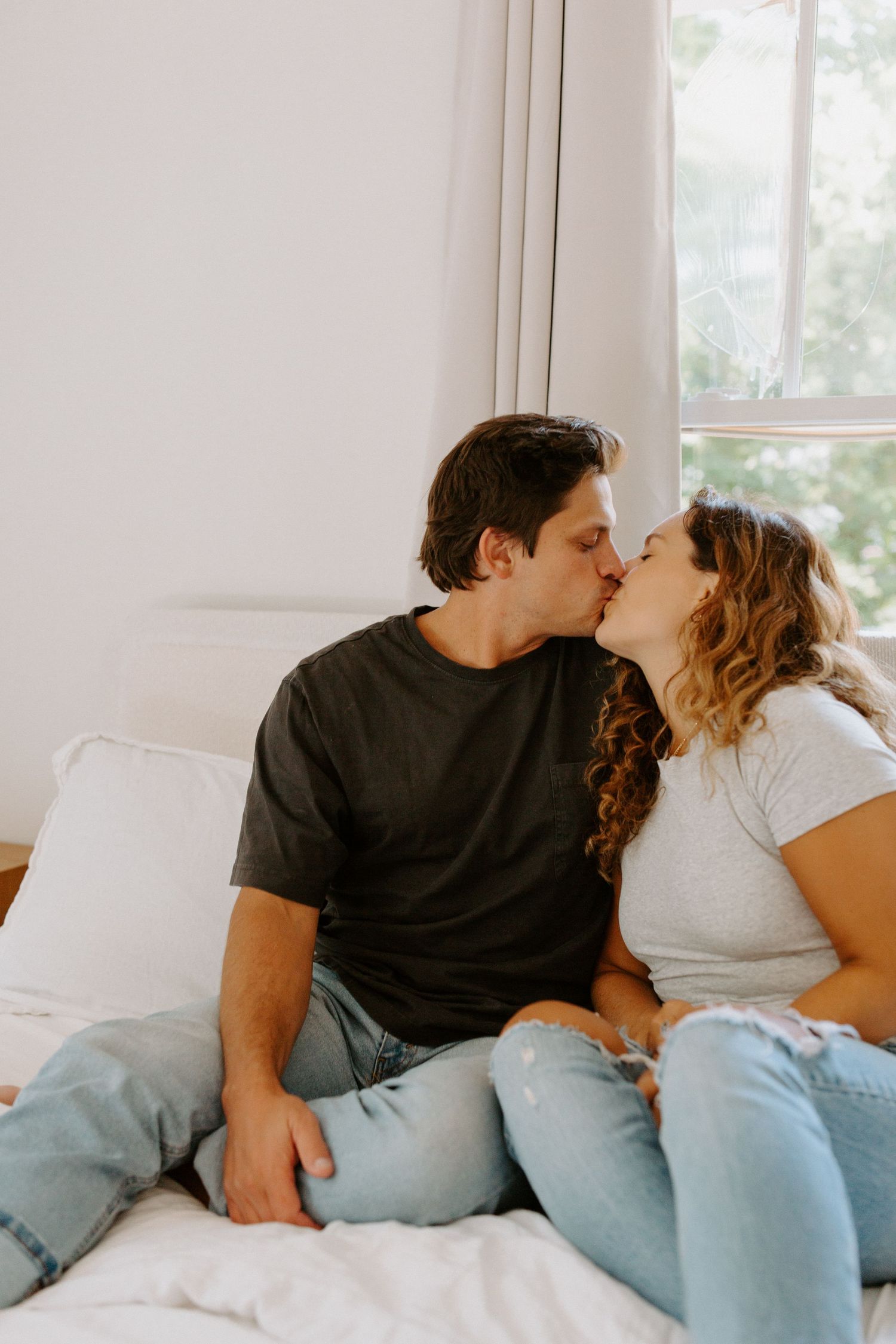 A couple shares an intimate moment while relaxing together on a white couch in a bright sunlit room.