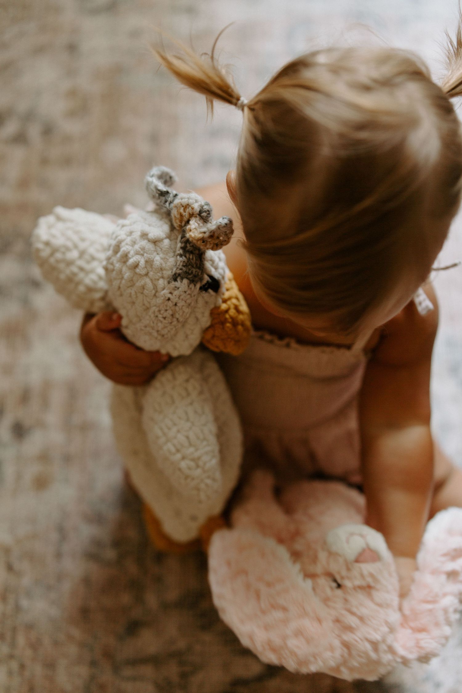 A toddler in brown clothing holds a white plush toy while sitting on a carpet.