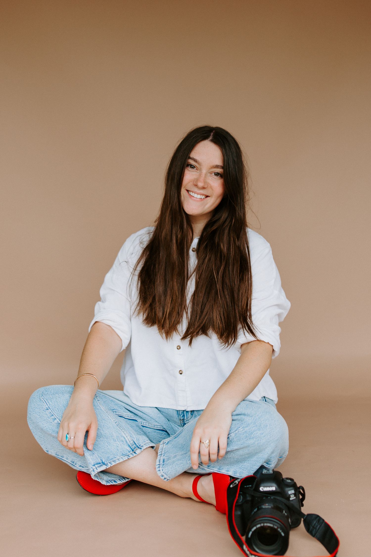 Studio photographer in white shirt and jeans poses with camera against brown backdrop.