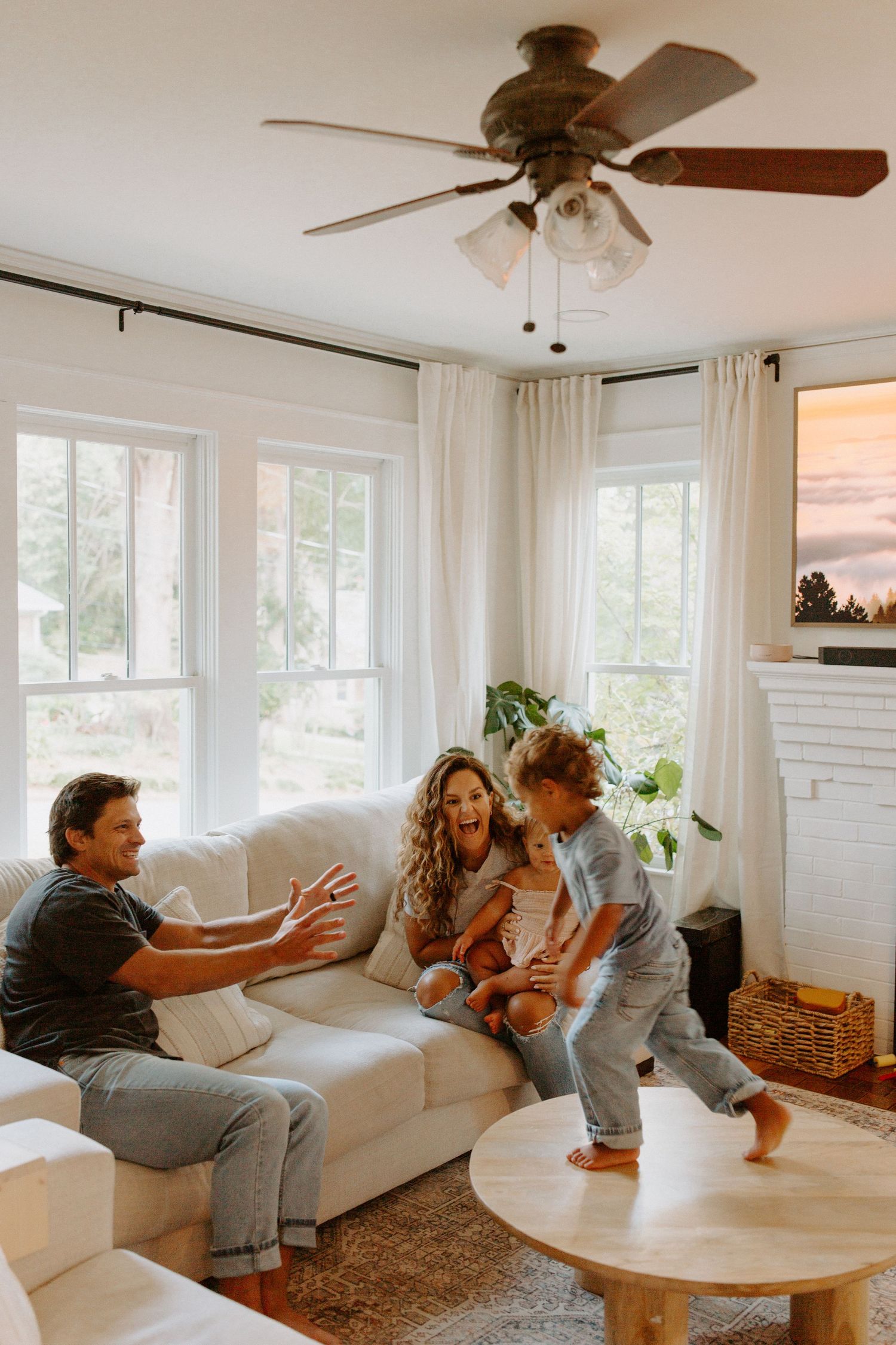 Family members play together on a white couch in a bright sunlit living room with large windows.
