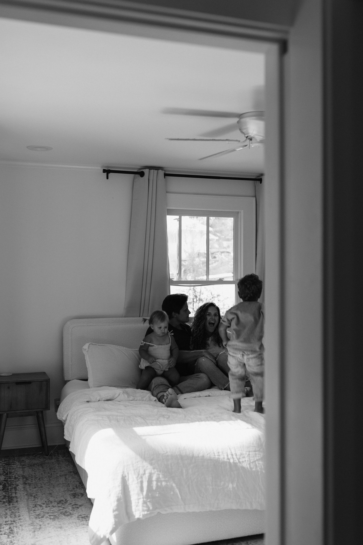 Black and white photo showing intimate family moments in a sunlit bedroom.