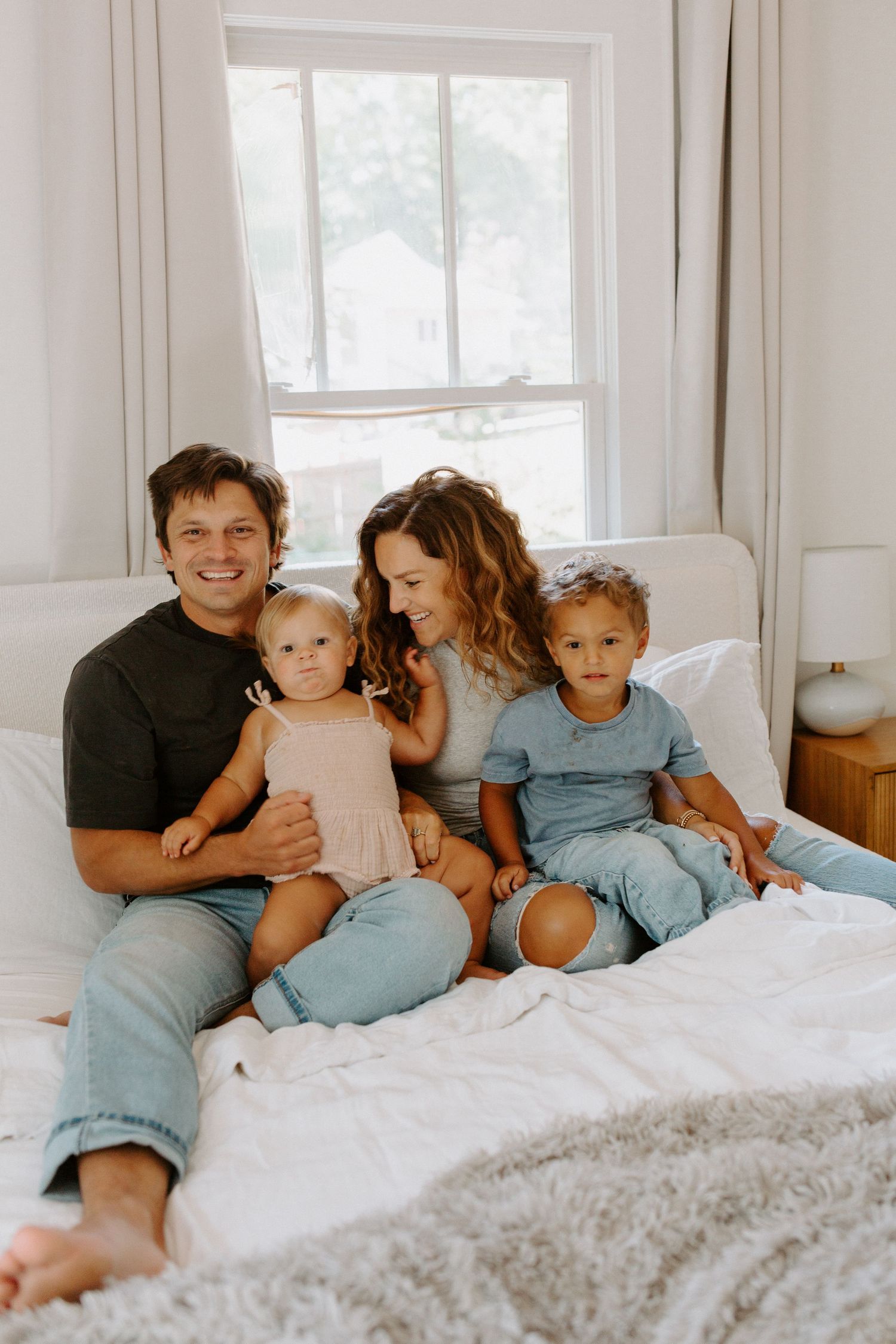 A casual family portrait on a comfortable bed with white linens and natural window light.