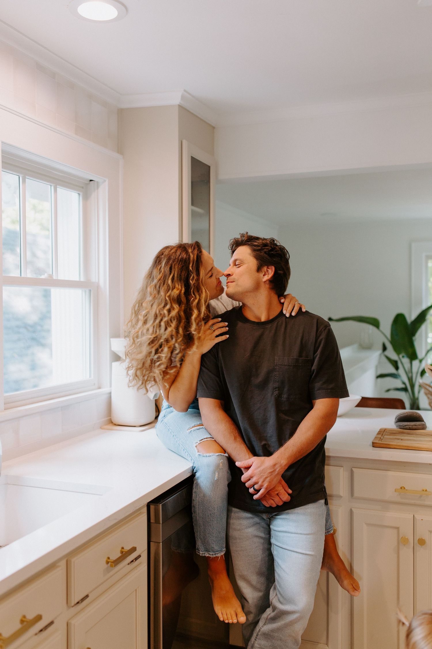 A couple sharing an intimate moment in a modern white kitchen with large windows.