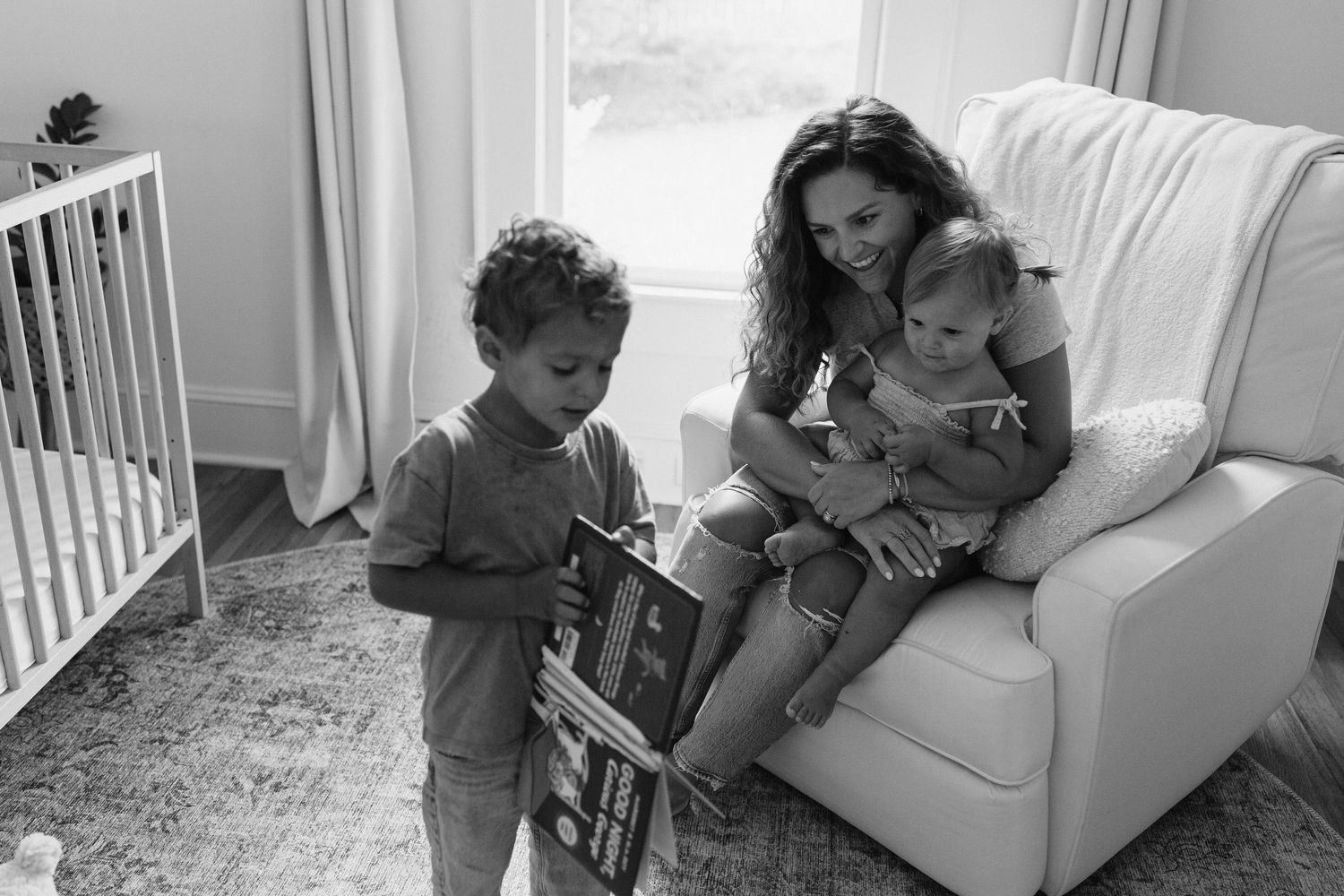 A black and white photo of a family reading together on a couch next to a baby crib in a nursery.